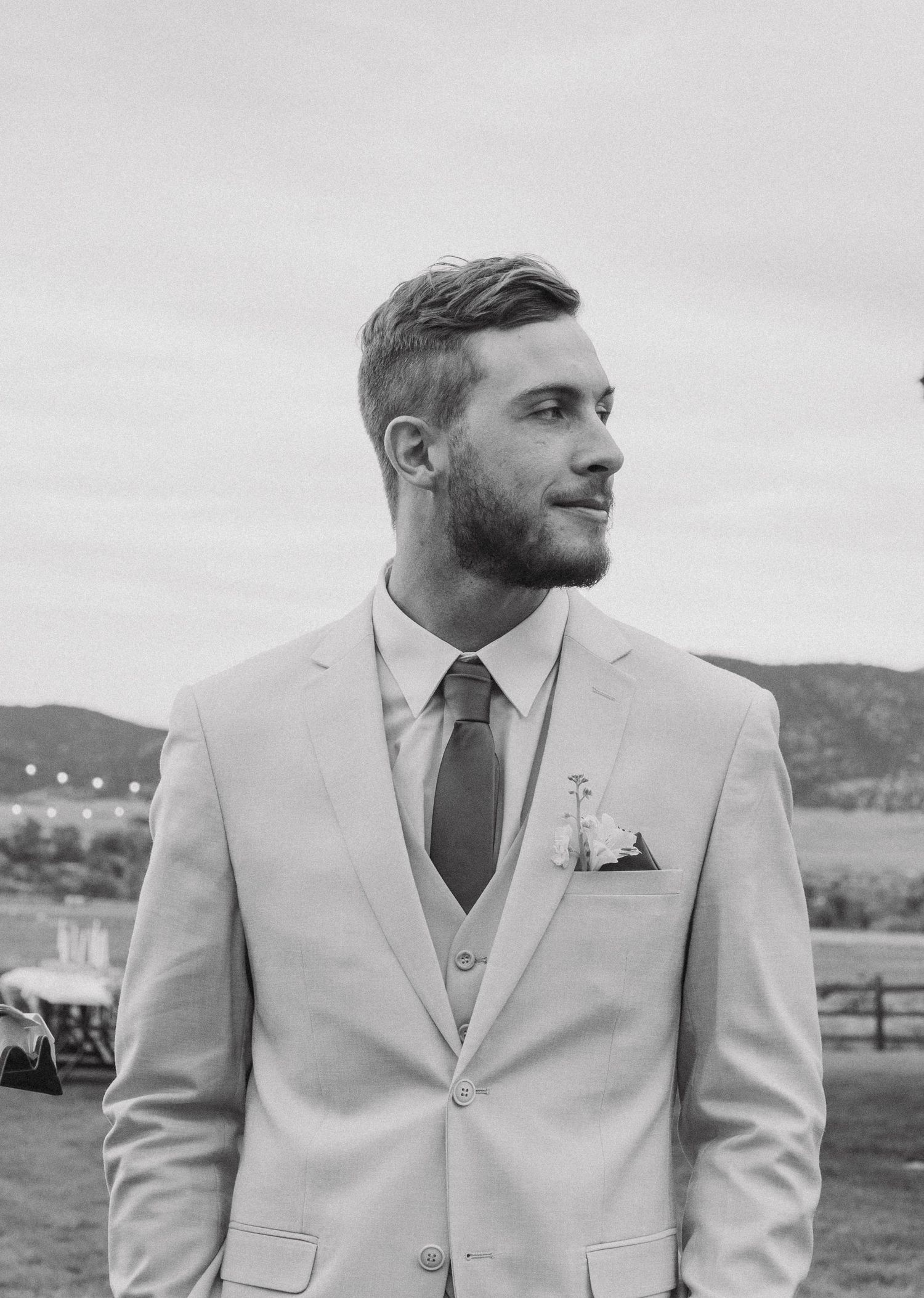 Groom in light beige three-piece suit poses against mountain landscape.