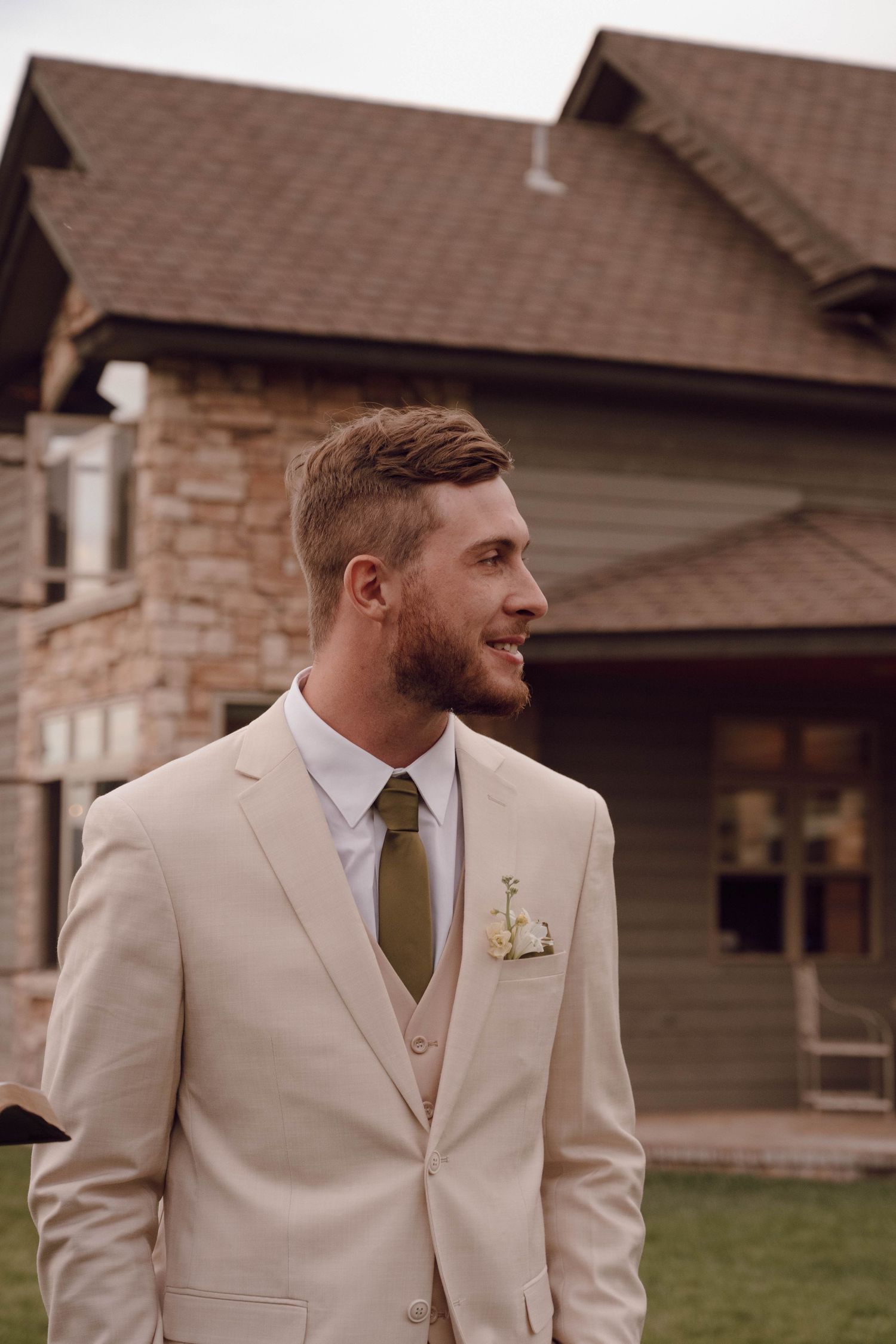A groom in a light beige suit and tie stands outdoors near stone buildings.