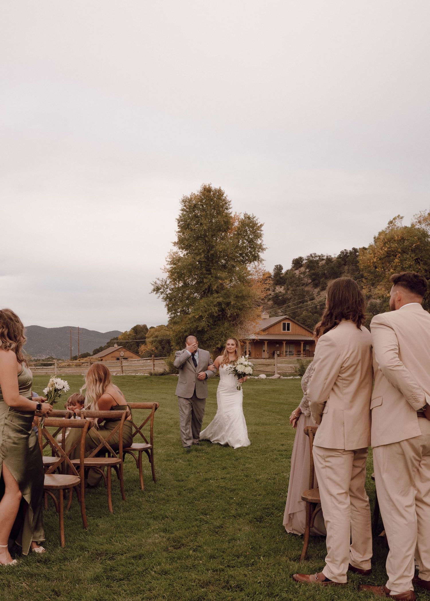 An outdoor wedding ceremony setup with guests gathering on a grassy lawn against a mountain backdrop.