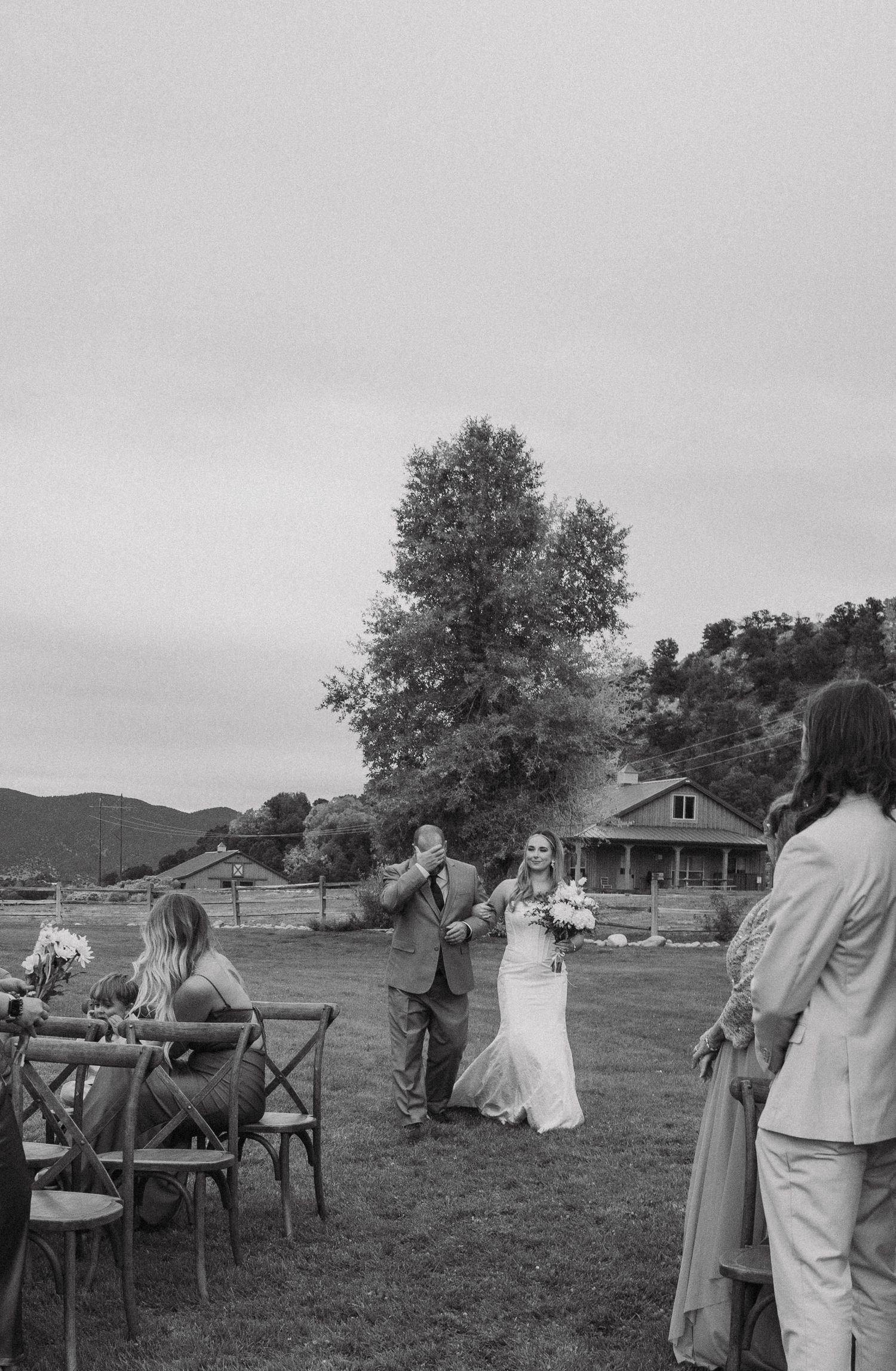 Black and white photo of wedding ceremony guests seated in chairs on grassy field with mountains in background.