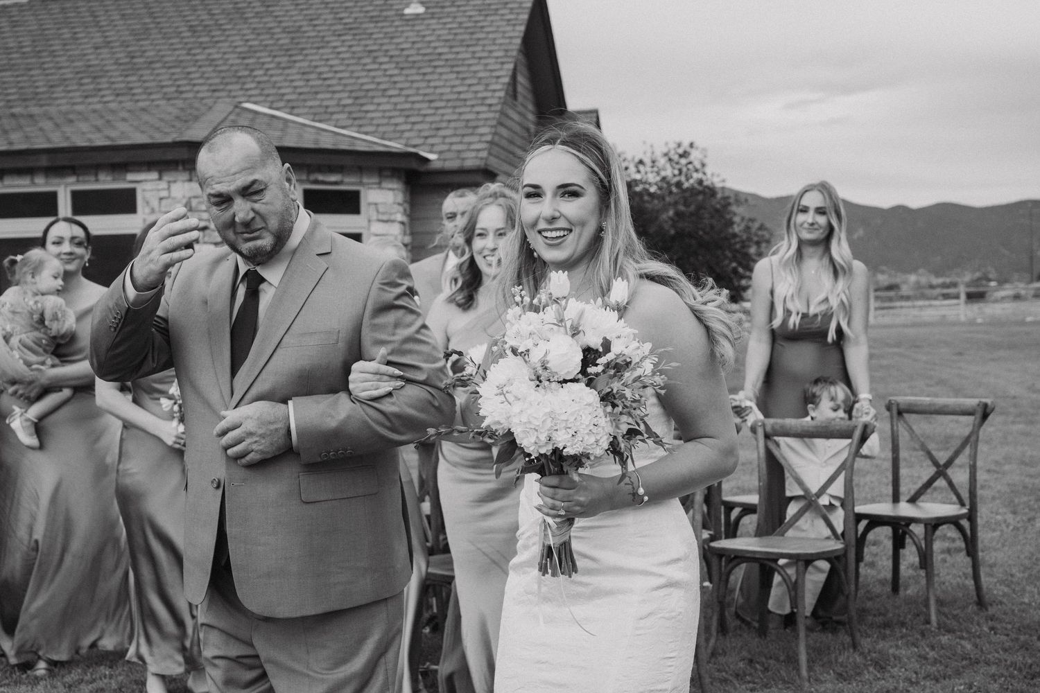 Wedding party poses for photos outdoors with mountains and rustic buildings in the background.