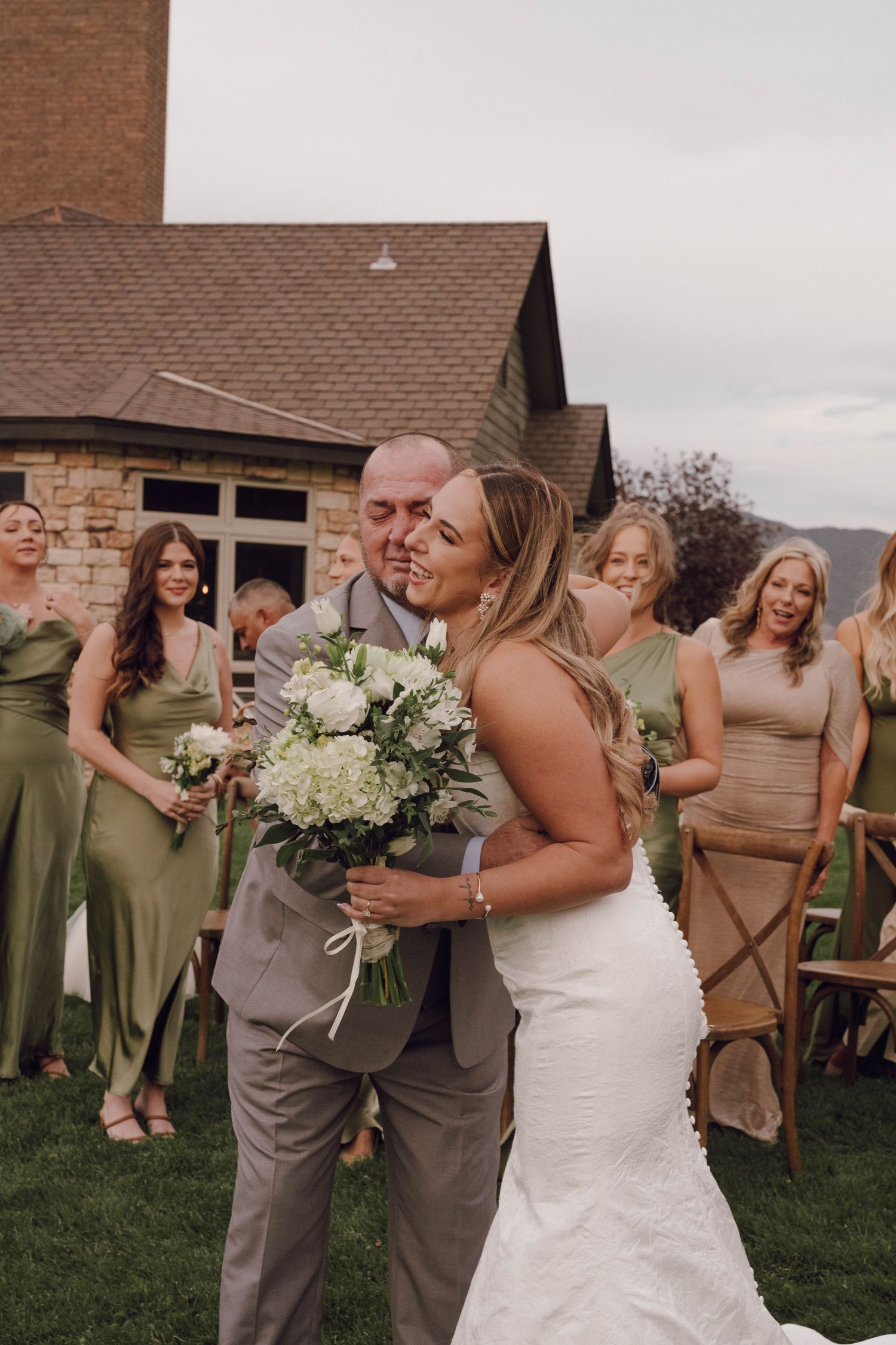 Wedding party celebrates outdoors with bridesmaids in olive green dresses and groomsmen in gray suits.