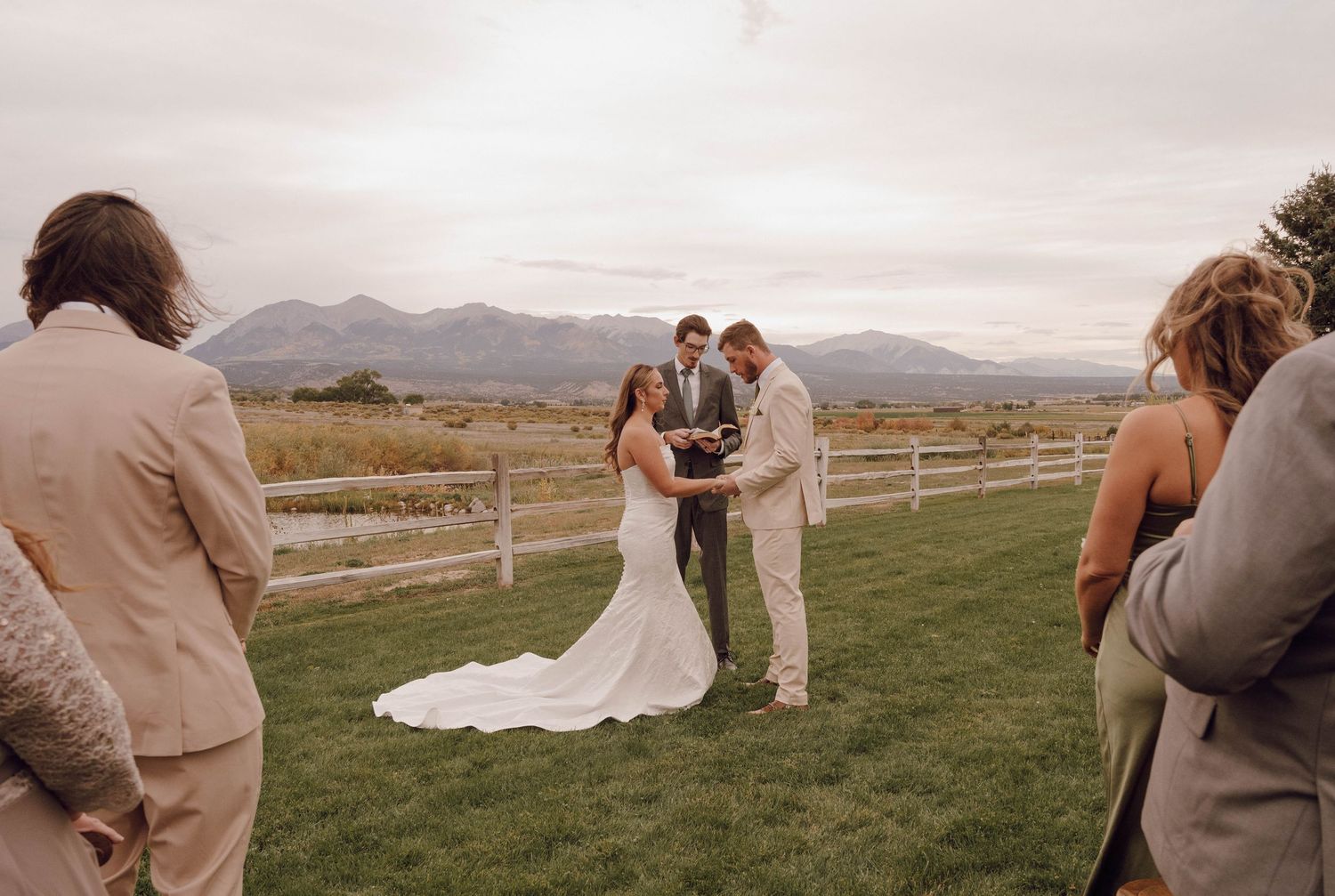 Wedding party gathered on grassy hillside with mountain landscape in background.
