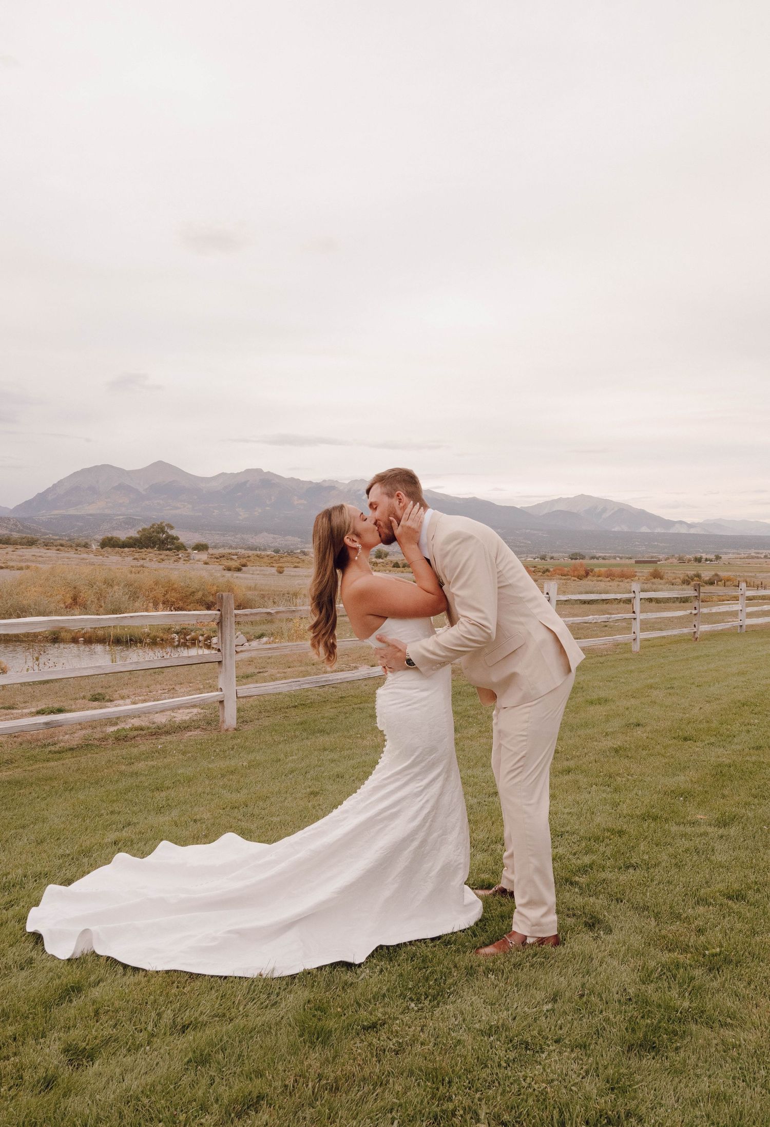 Romantic wedding couple poses in front of mountain landscape with white fence and flowing dress.