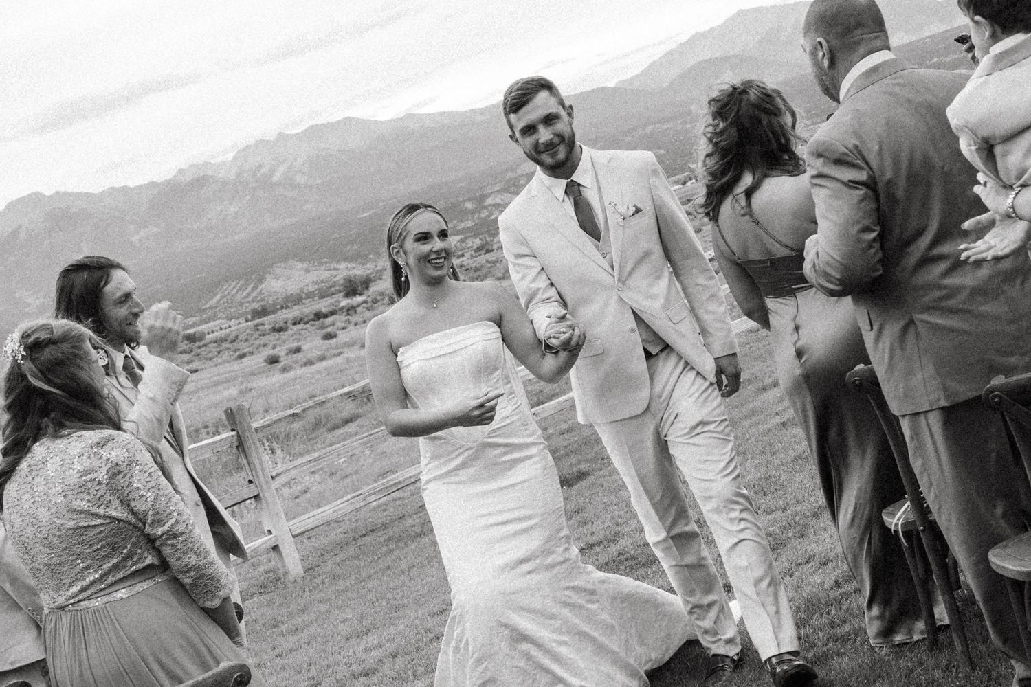 A joyful wedding party celebrates outdoors with mountains visible in the background.