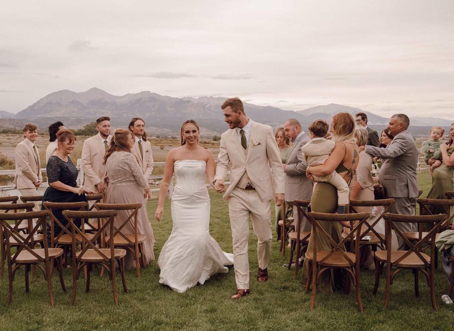 Wedding celebration on a grassy hillside with rustic wooden chairs and mountain scenery.