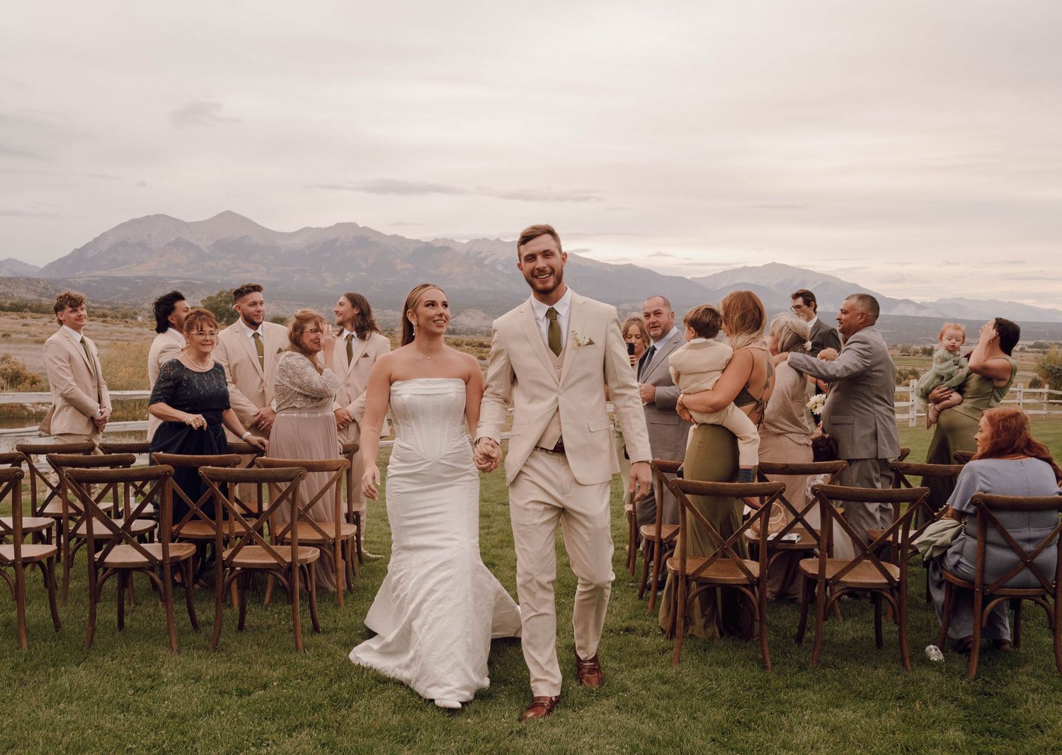 Guests celebrate at an outdoor wedding reception with mountain views and wooden cross-back chairs.