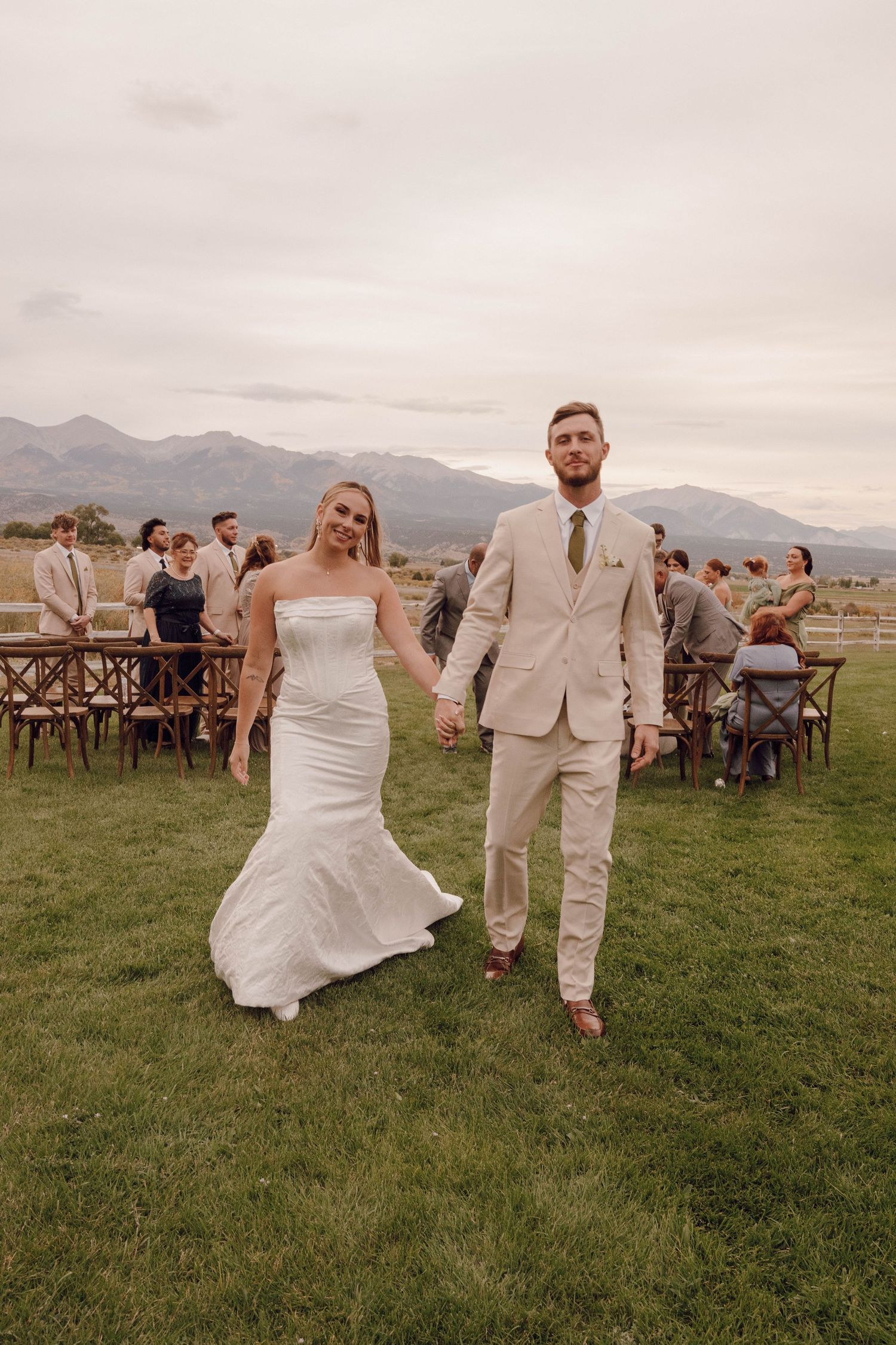 Couple walks across grassy field during outdoor wedding reception with mountain backdrop and rustic seating.