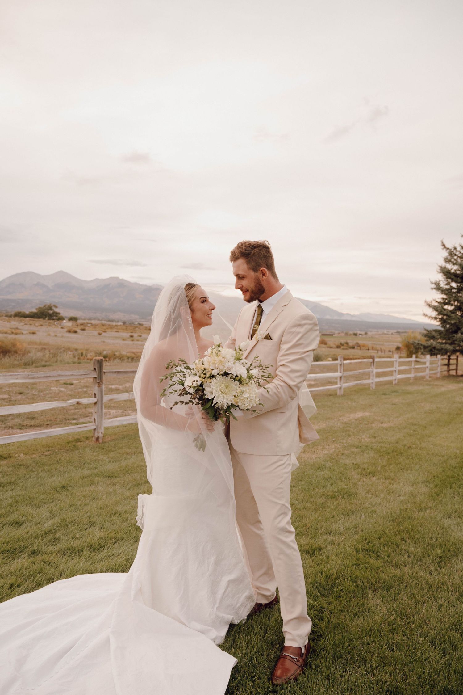 Couple in white and beige share romantic moment on grassy hill with mountain backdrop.