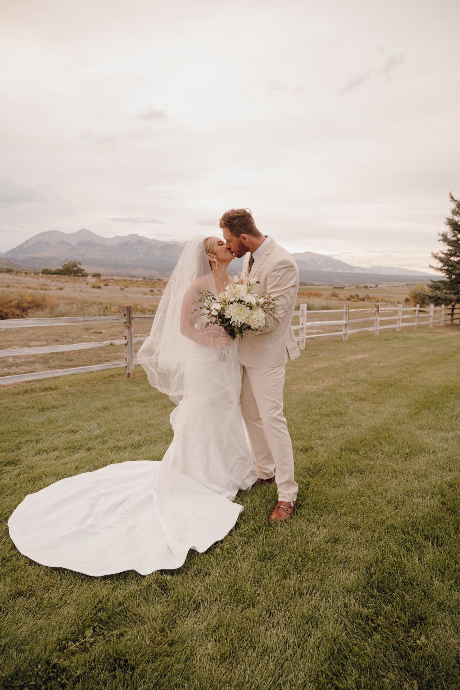 Newlyweds share romantic moment in field with mountain backdrop and flowing white dress.