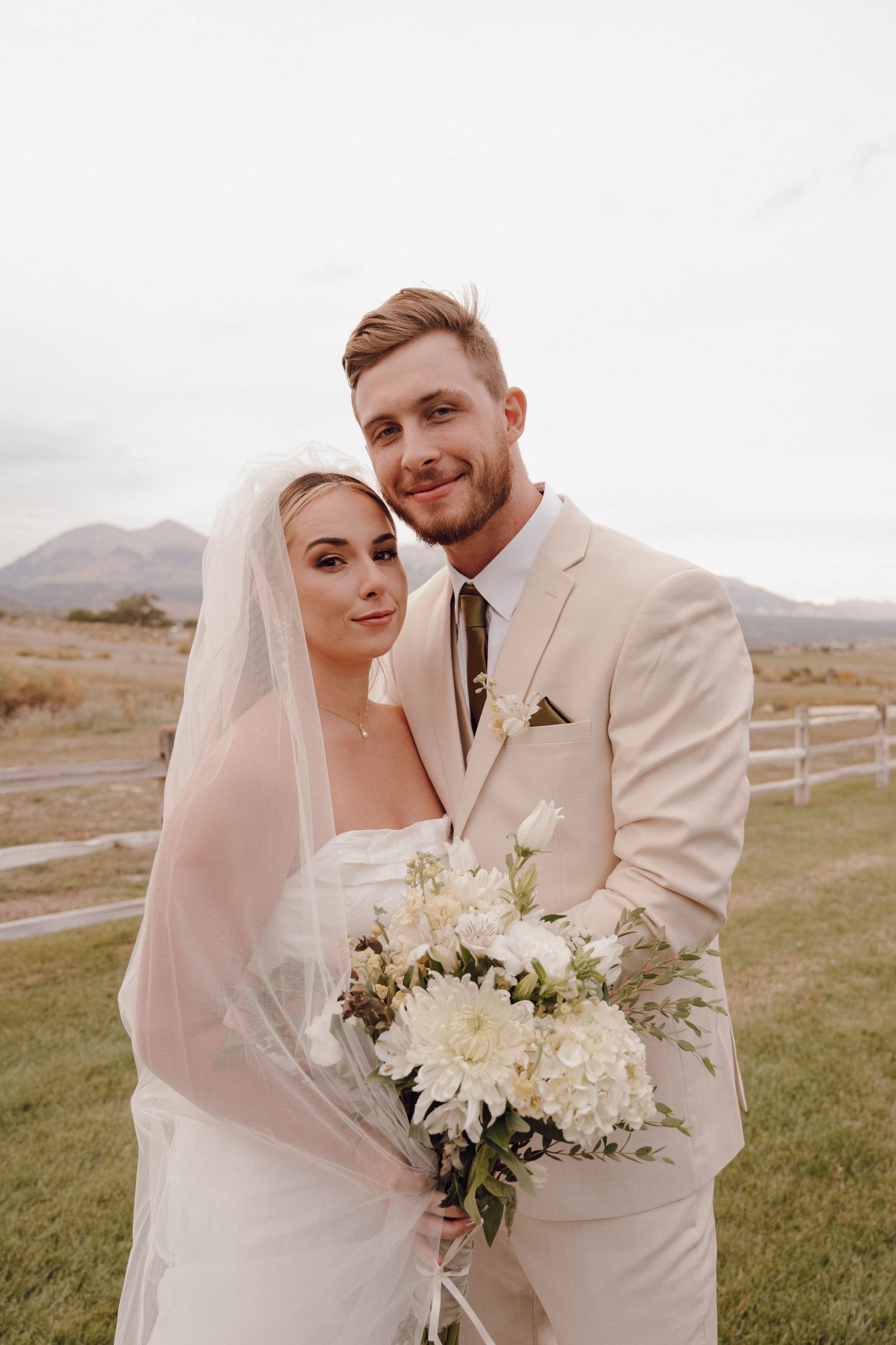 A romantic wedding portrait of a couple in cream attire embracing against a mountain backdrop.