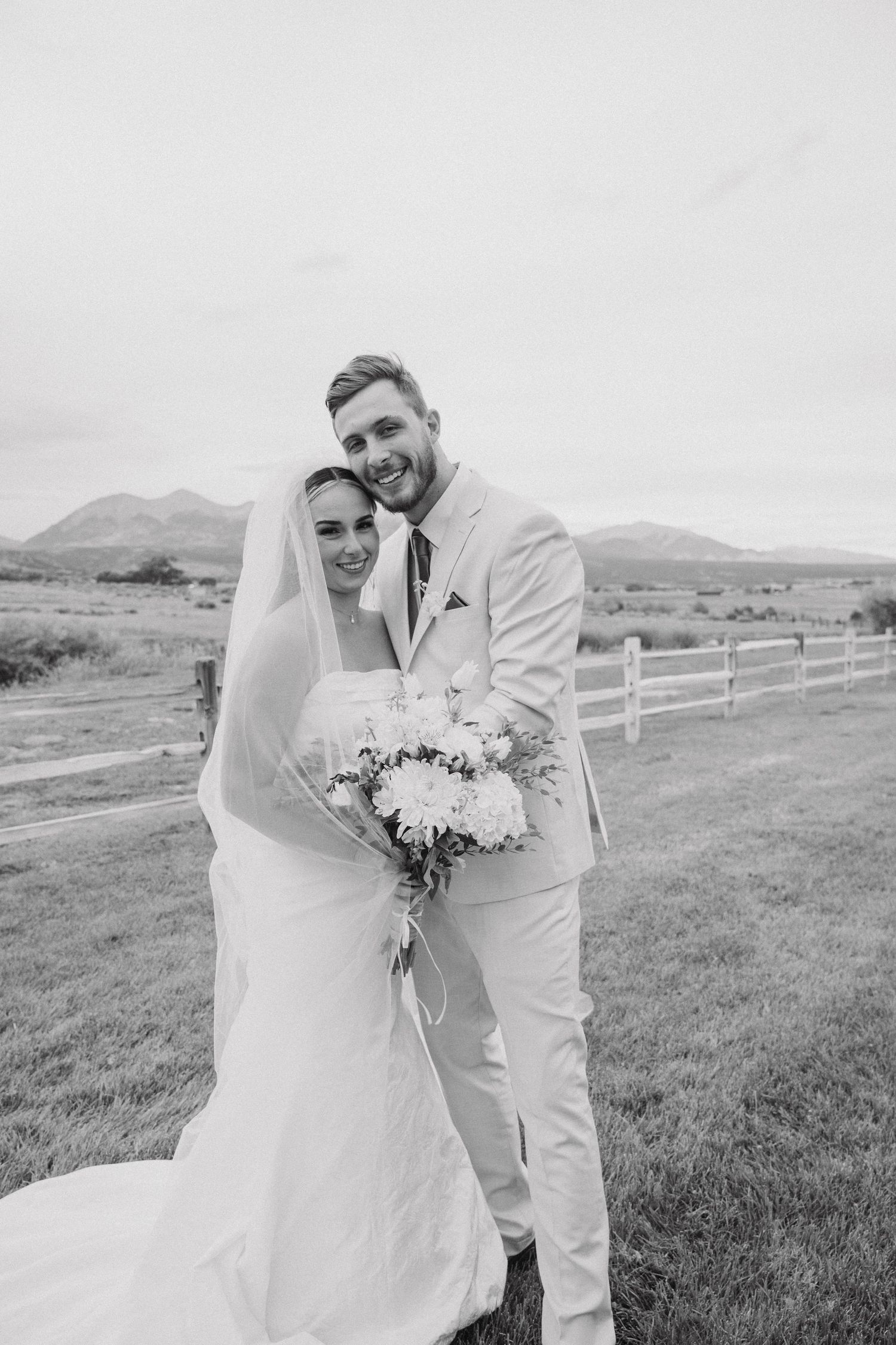 Black and white wedding photos show a couple embracing in a scenic mountain field with white fencing.