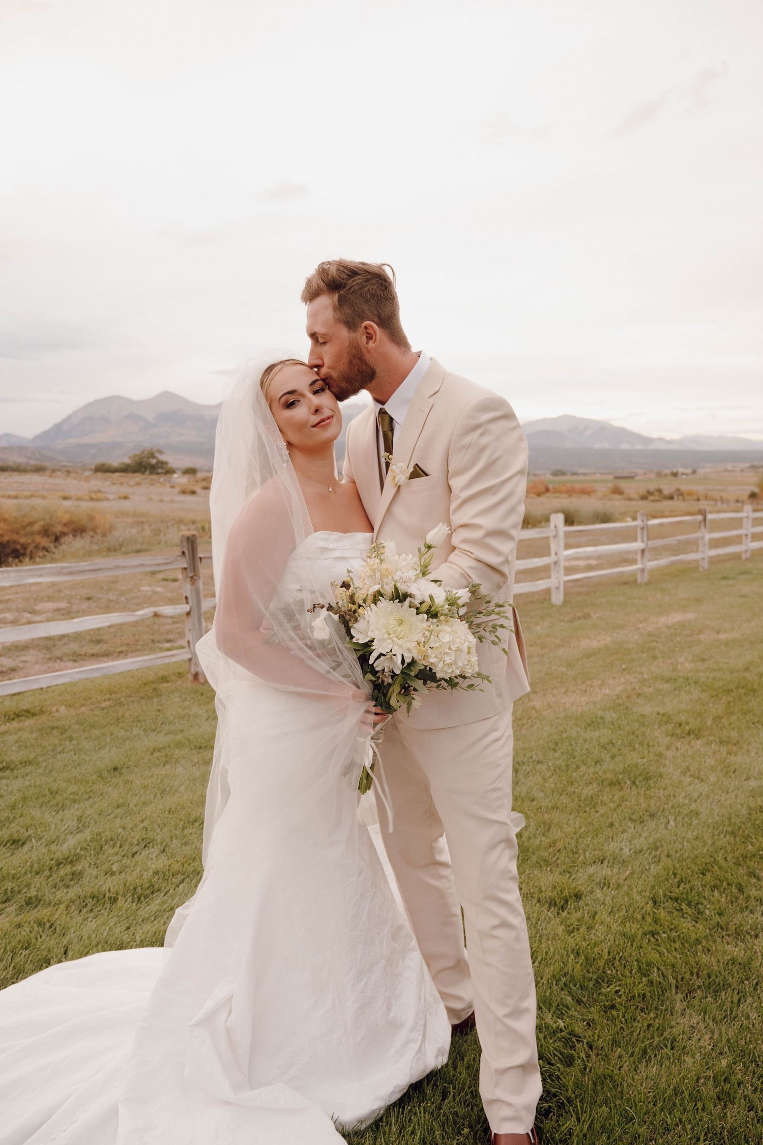 A romantic wedding photoshoot in a scenic mountain landscape with white fencing and flowing veil in the breeze.