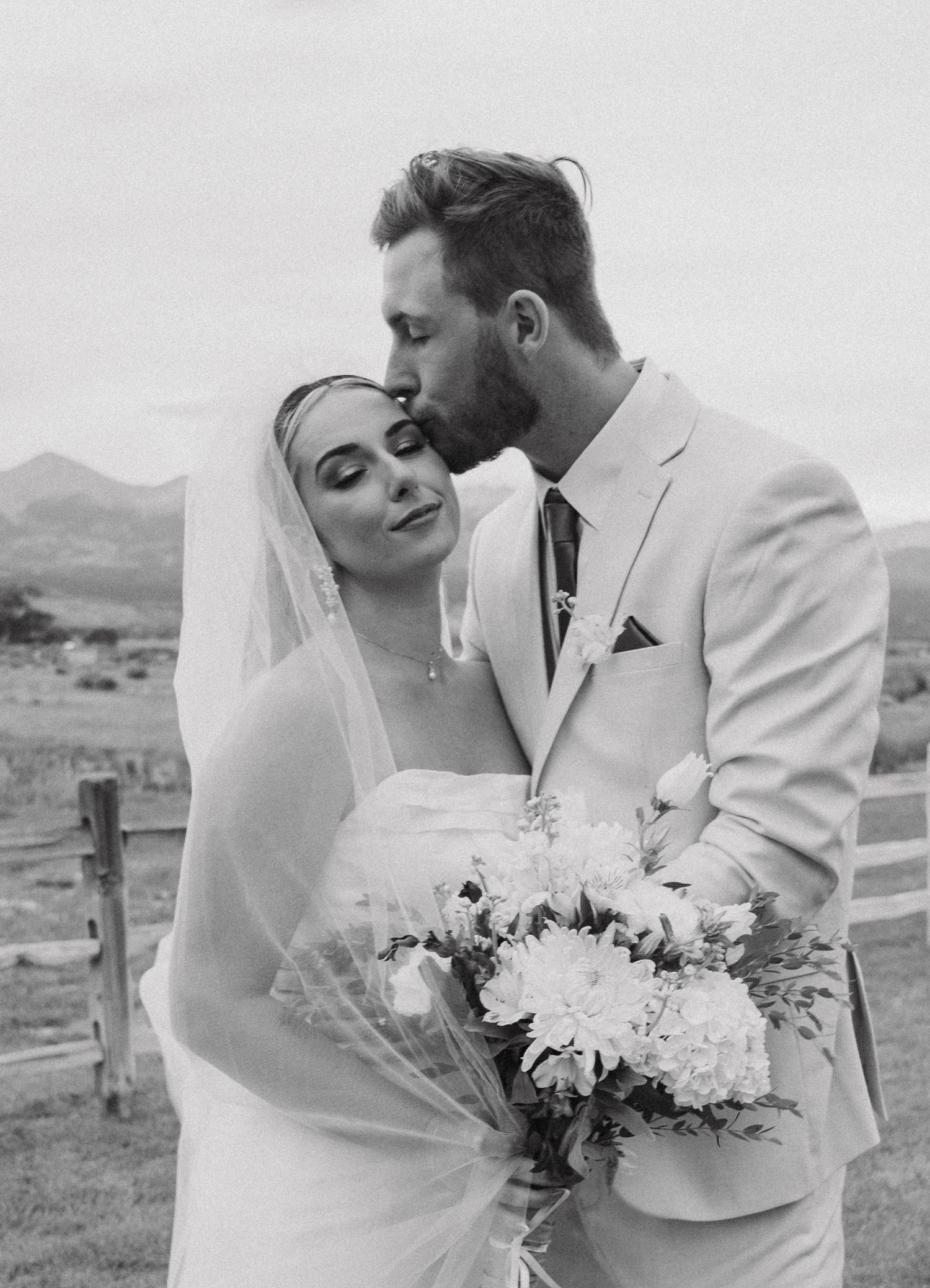 A romantic black and white wedding photo showing a tender moment between newlyweds with mountains in the background.