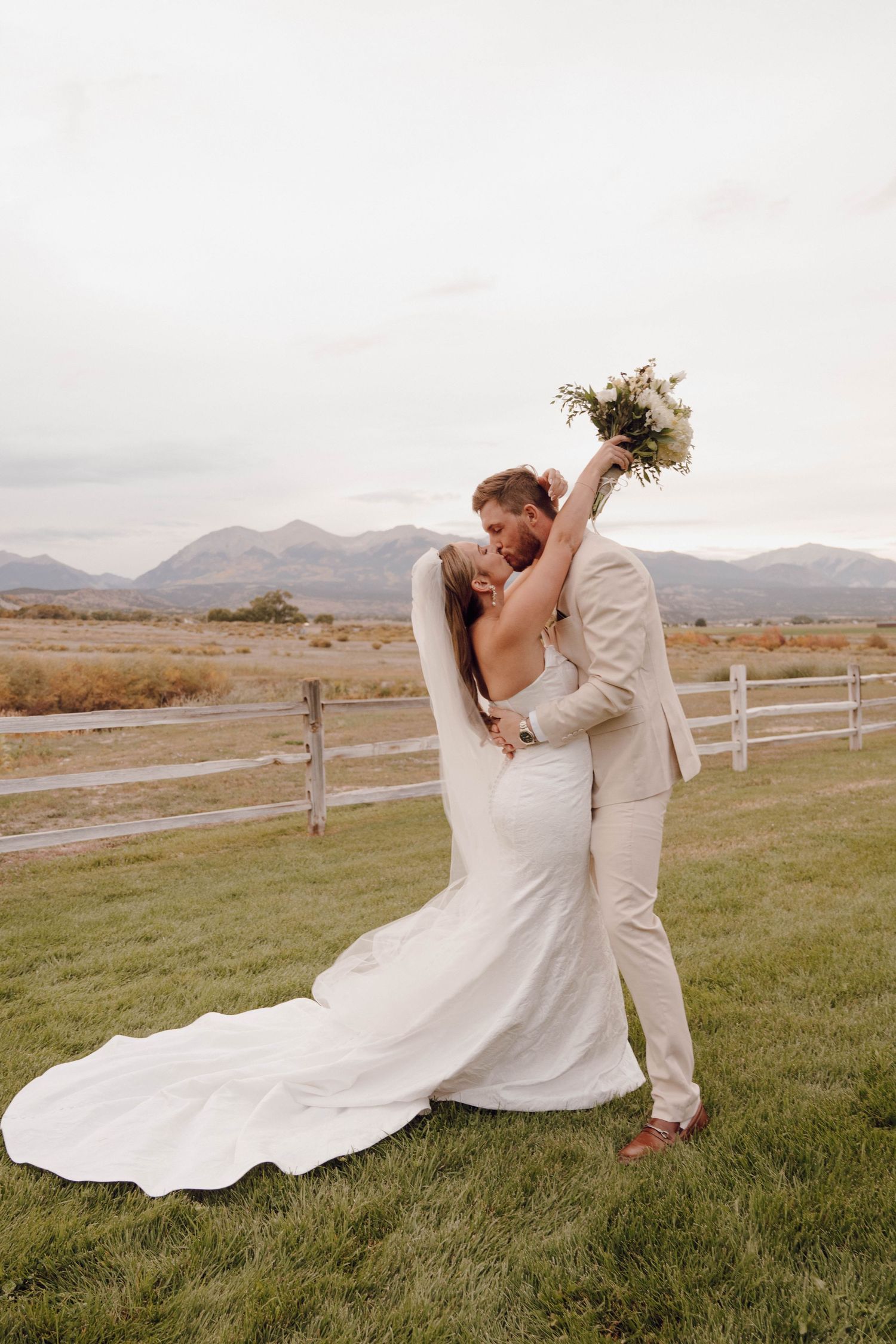 Wedding couple in white and cream sharing intimate moments on grassy field with mountains in background.