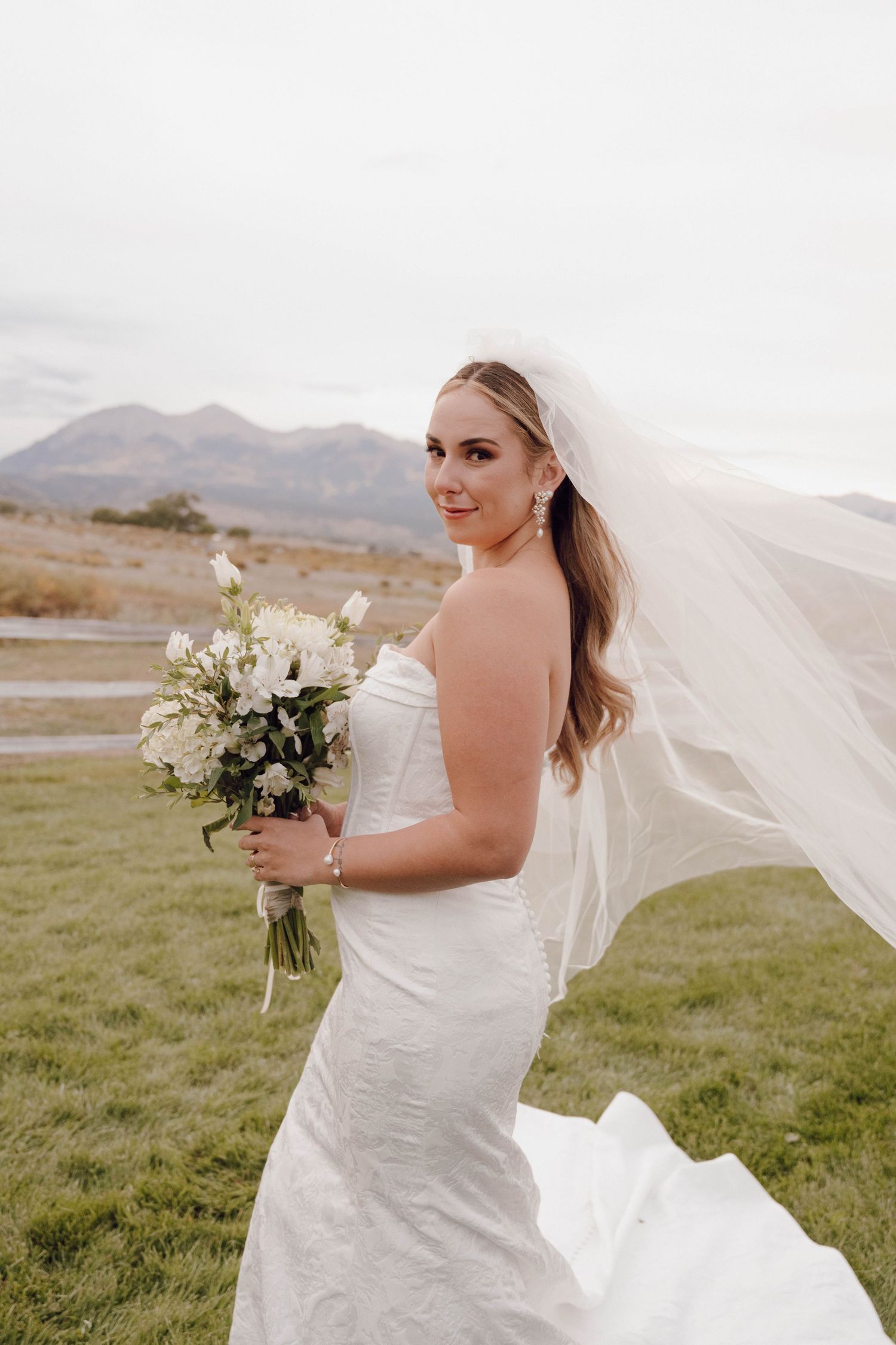 Bride in white wedding dress poses with flowing veil against mountain backdrop.
