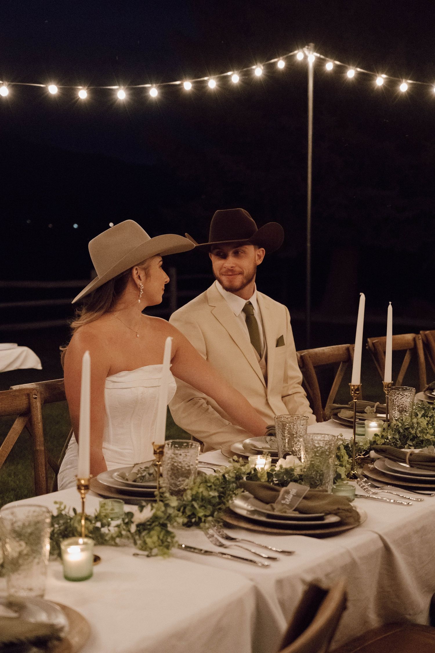 Wedding reception table under string lights with elegant place settings and greenery decor.