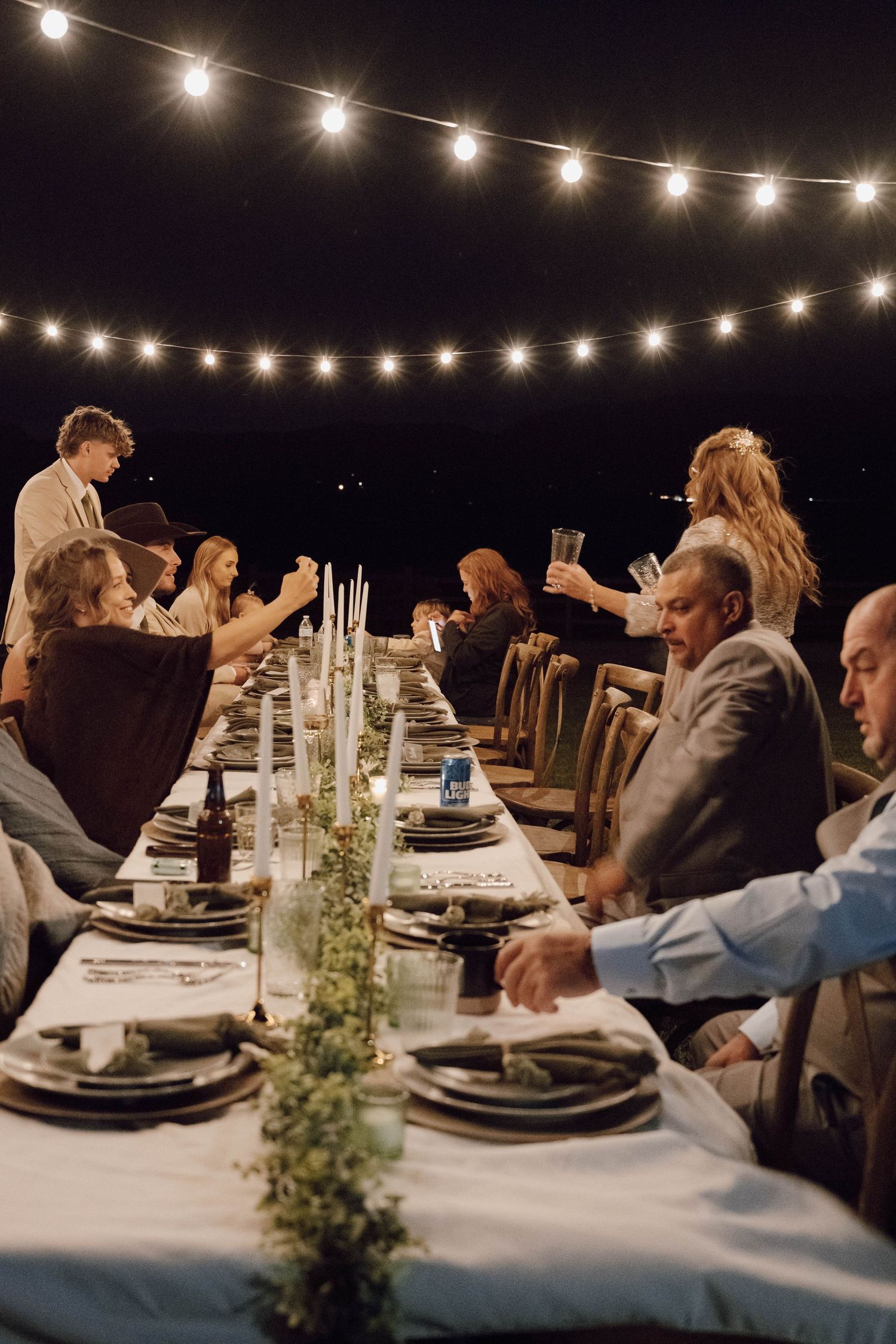 People gather for an elegant outdoor dinner party under string lights with a long table set with candles and greenery garland.