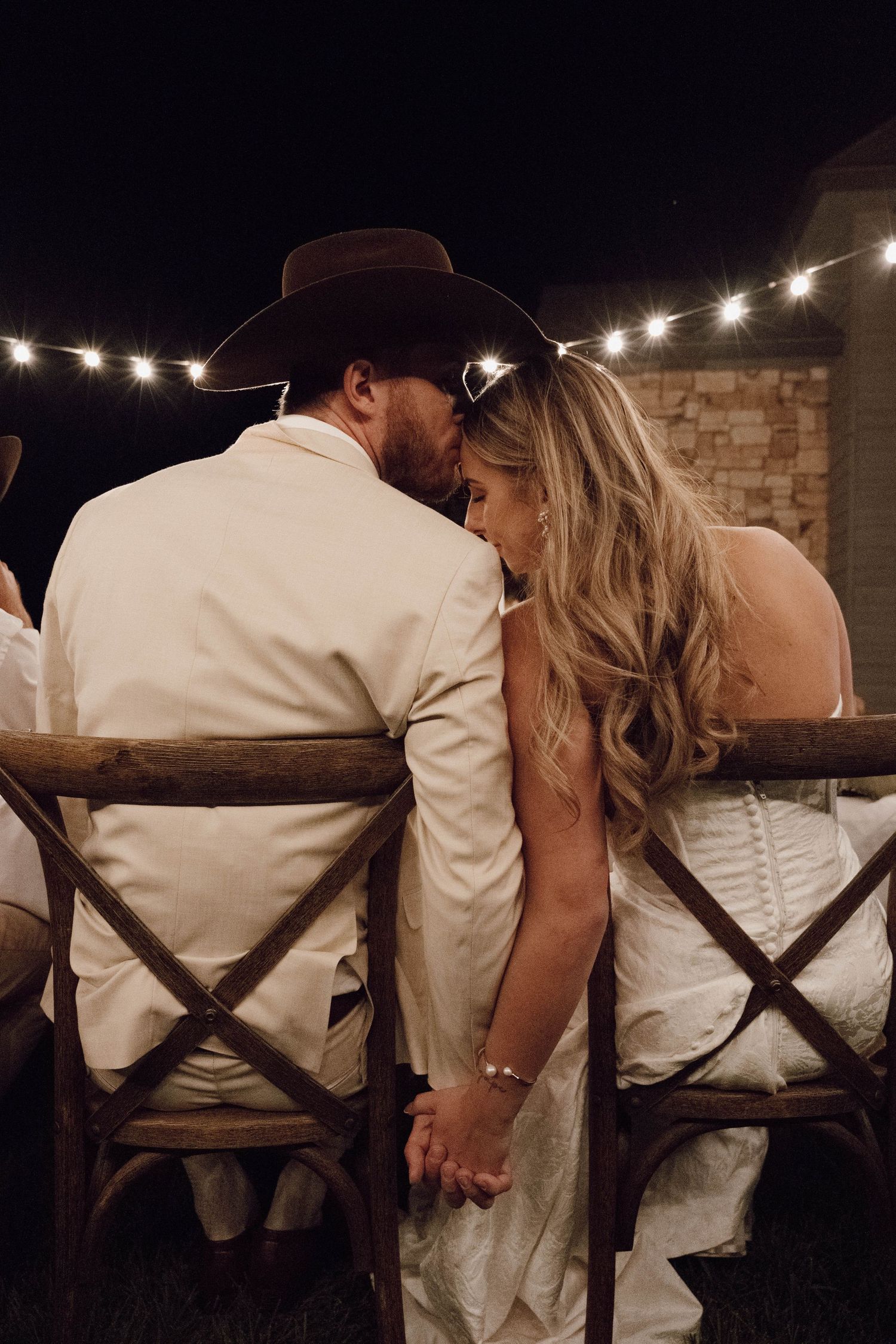 Romantic evening scene of couple sitting close together under string lights at outdoor reception.