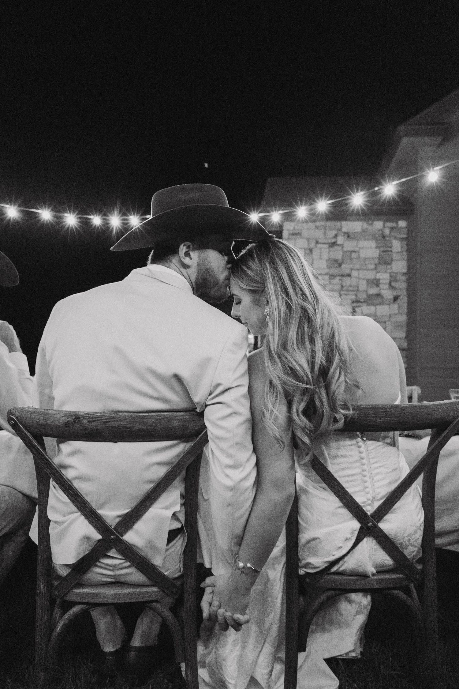 Wedding party seated in director chairs showing backs with X pattern during outdoor evening reception.