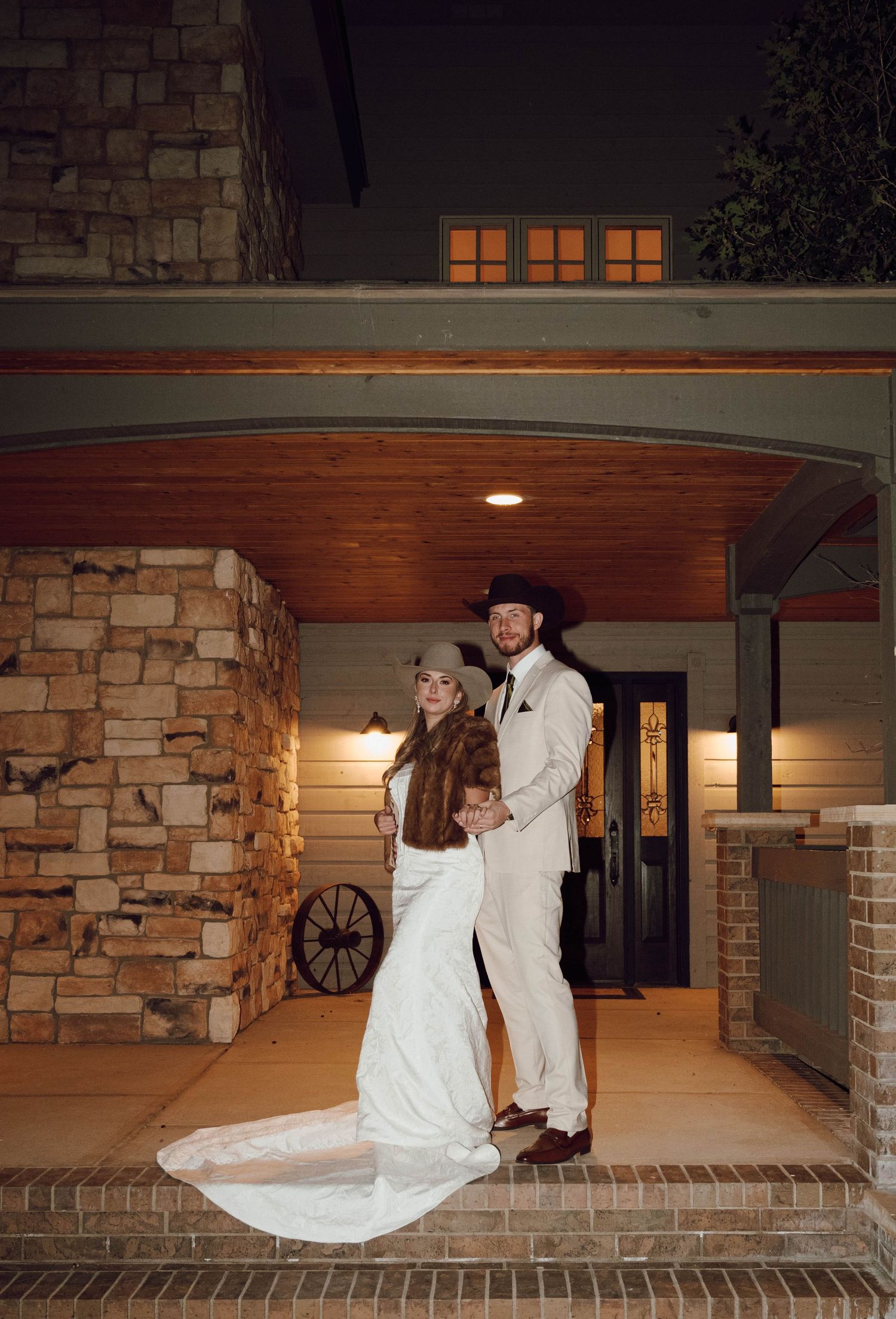 A couple in wedding attire poses for romantic photos on a brick porch with stone columns at night.