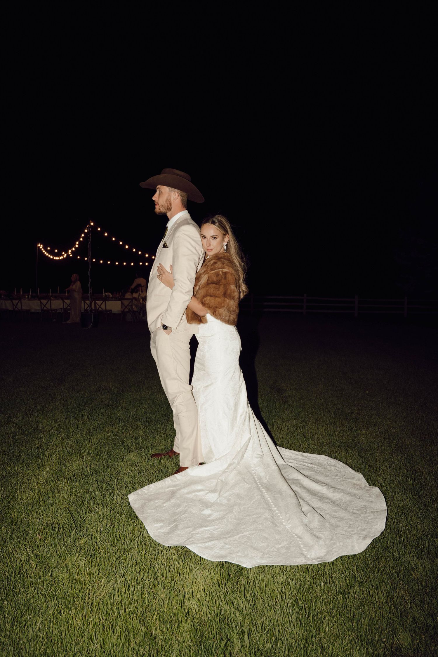 Nighttime wedding photo of couple dancing on grass with string lights in background.