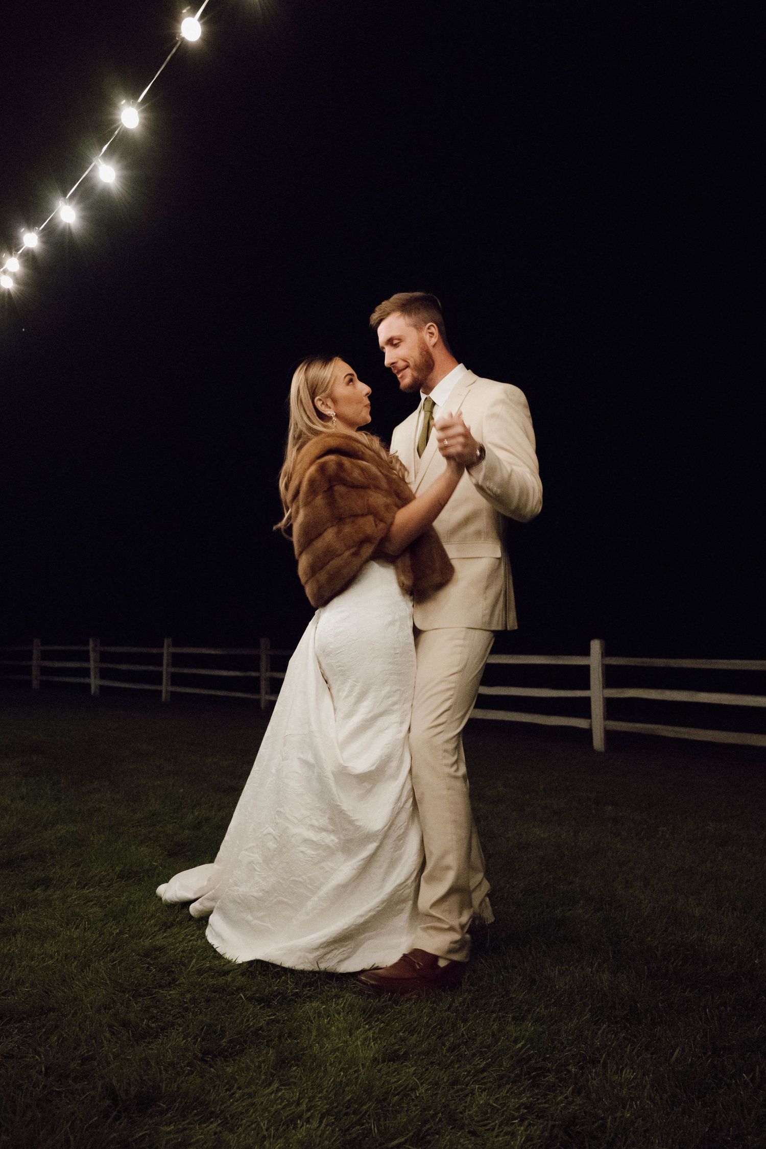 A romantic nighttime wedding dance under string lights with the bride wearing a fur wrap over her white gown.