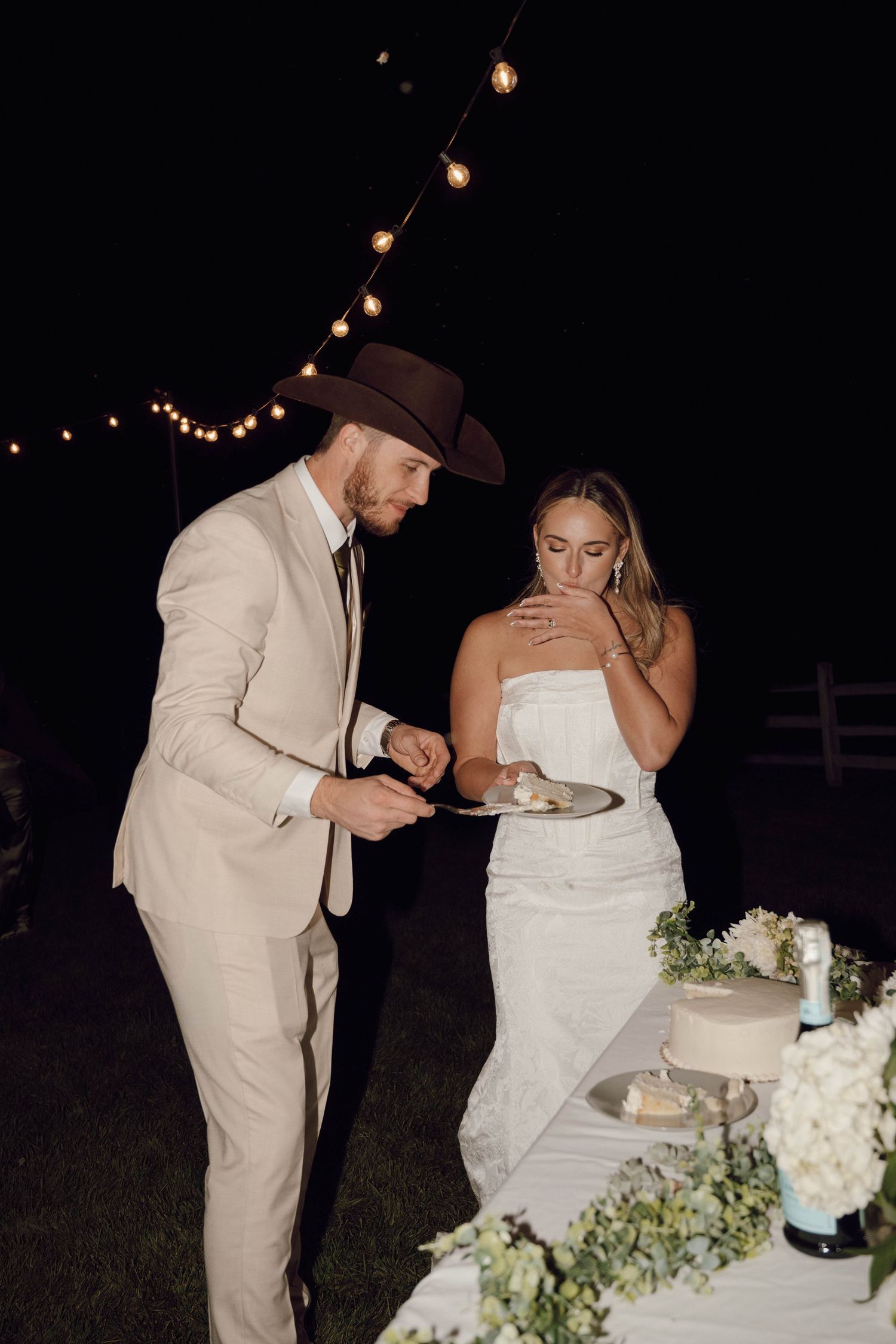 Newlyweds share a playful cake-cutting moment at their outdoor reception under string lights.