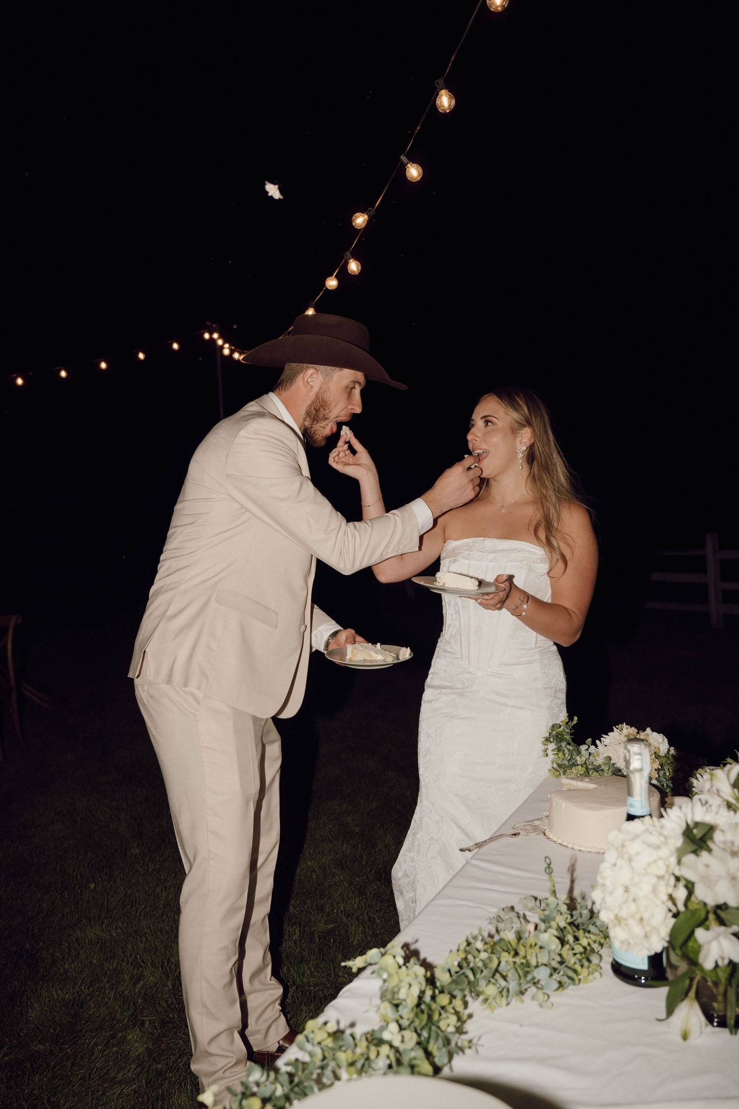 Newlyweds playfully feed each other wedding cake at an outdoor evening reception with string lights overhead.