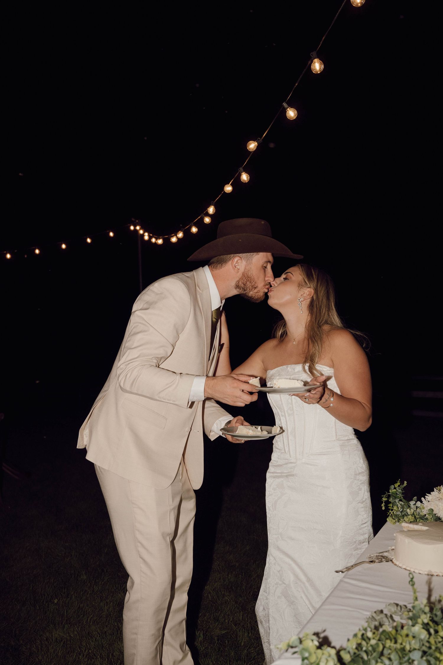 Newlyweds share cake during their wedding reception under twinkling string lights.