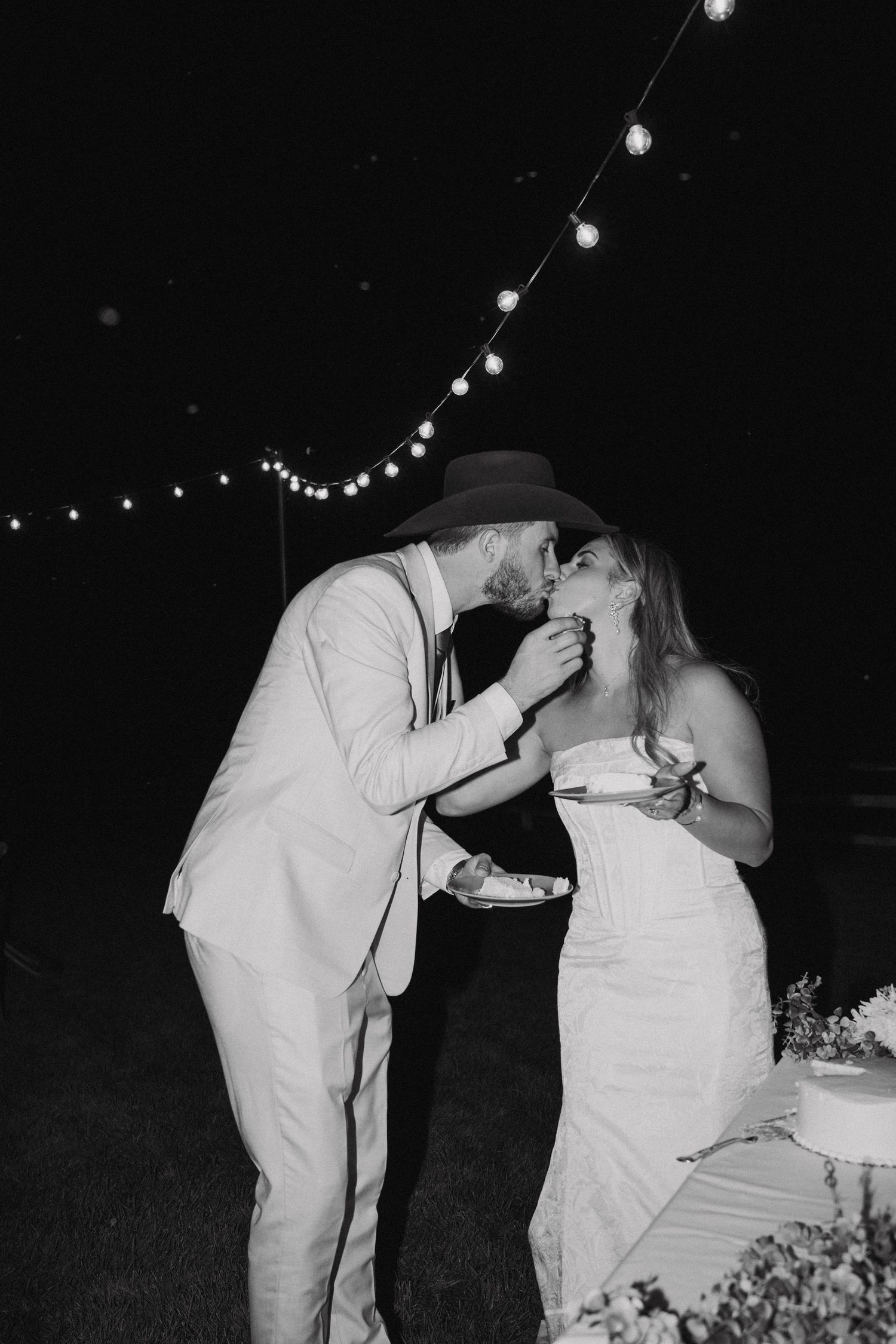 A romantic wedding couple sharing their first kiss at an outdoor evening reception under string lights.