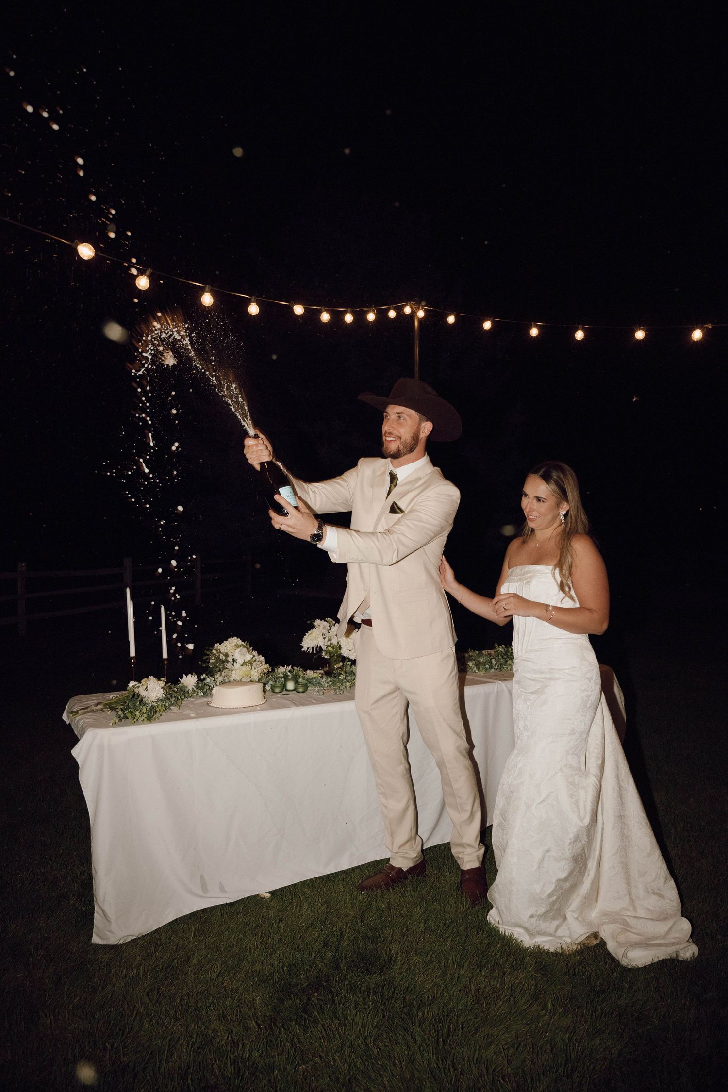Couple lighting sparklers at night during outdoor wedding reception with string lights overhead.