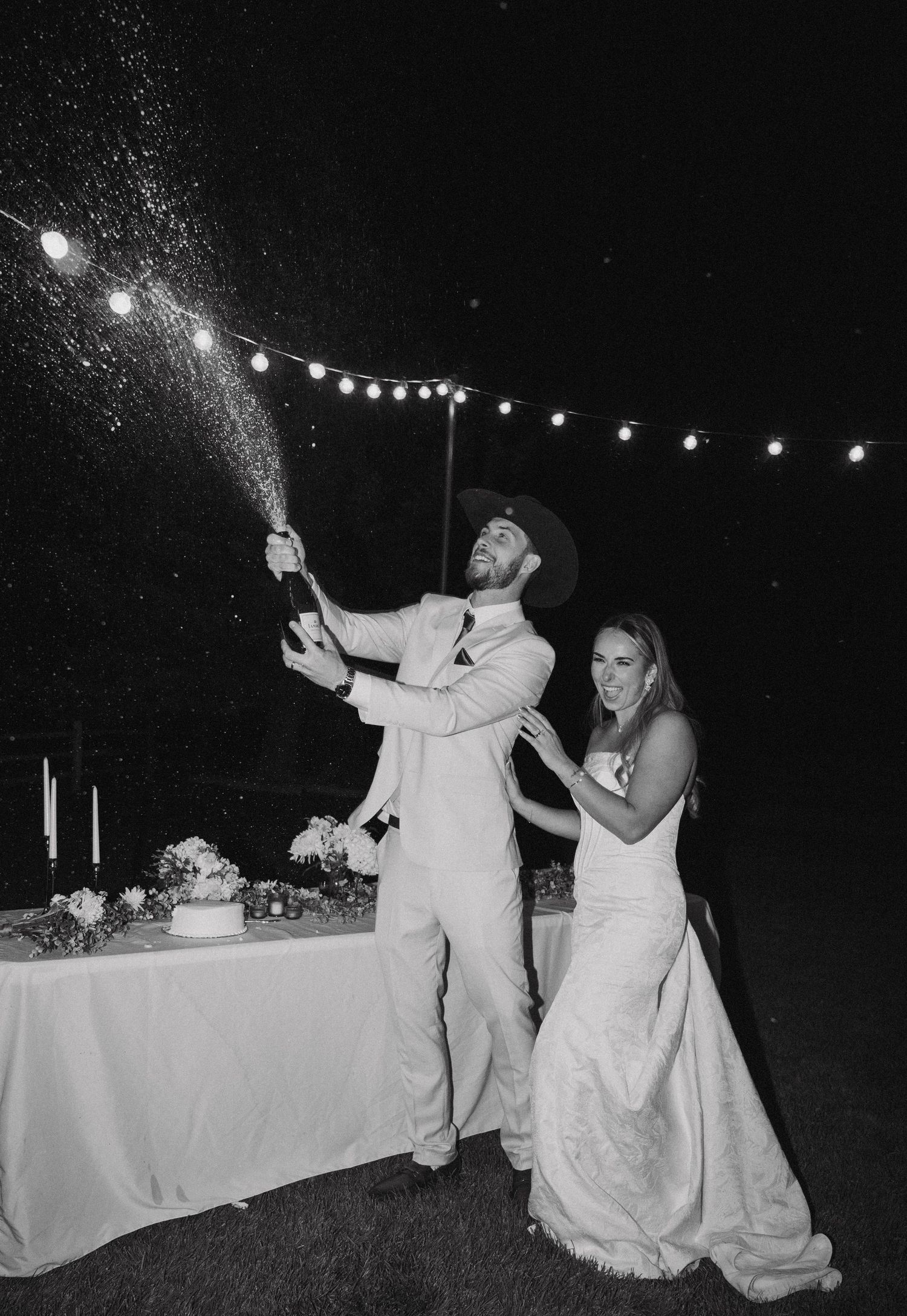 Wedding couple shares a playful moment at an outdoor evening reception with string lights overhead.