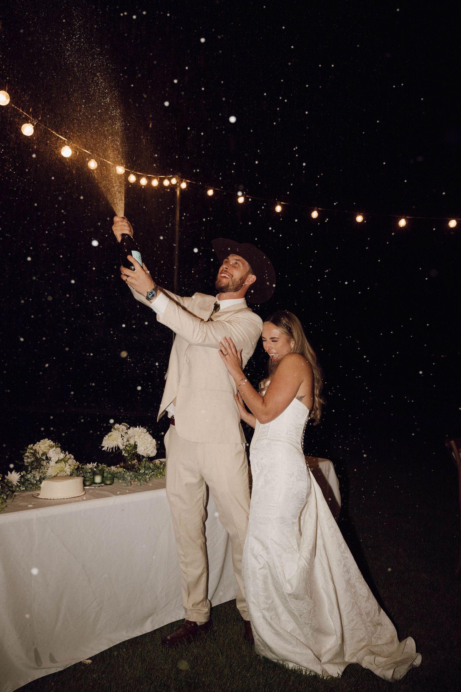 A couple celebrates under string lights at their outdoor nighttime wedding reception with falling snow.