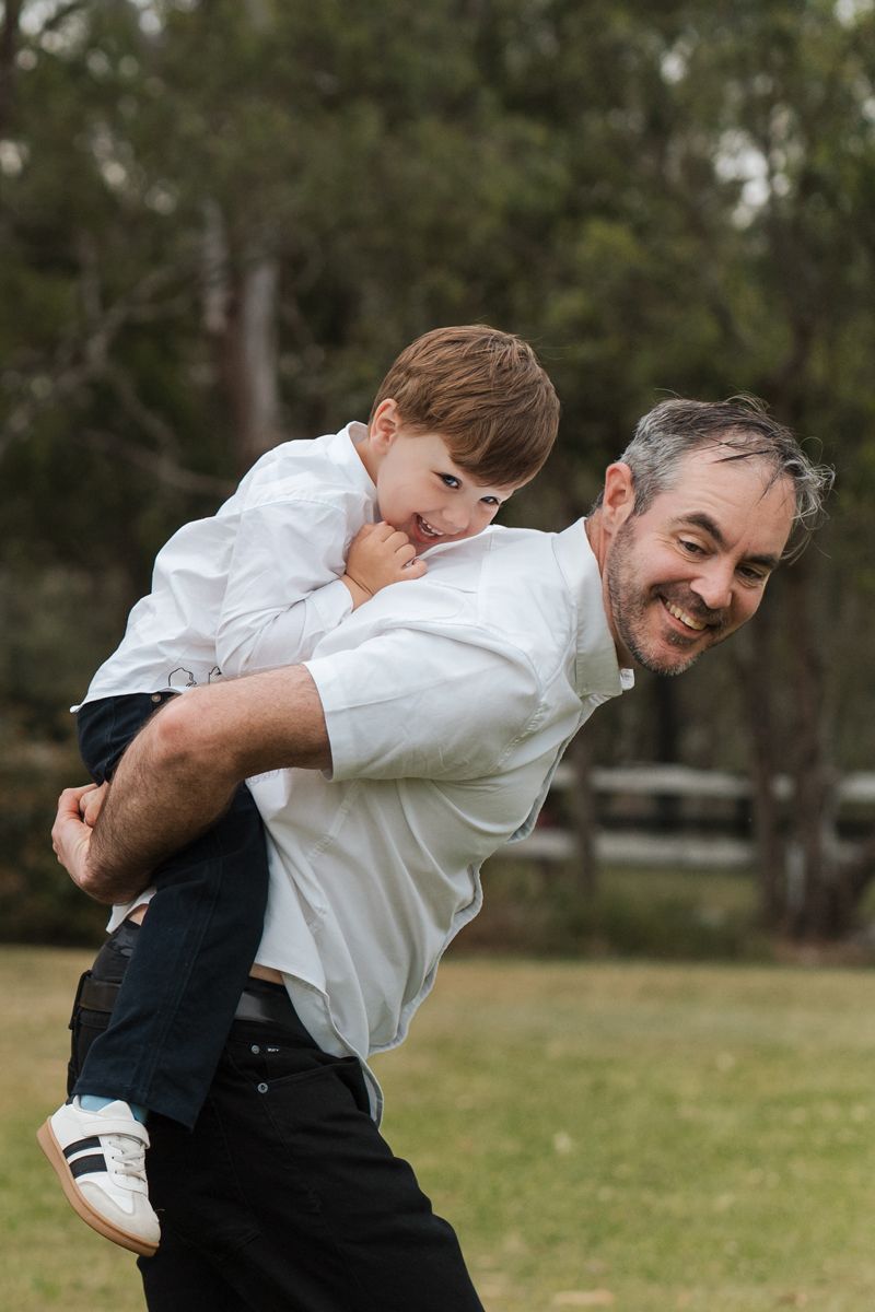 A playful moment outdoors as a young boy rides piggyback on someone wearing a white shirt against a wooded background in Sydney family photo