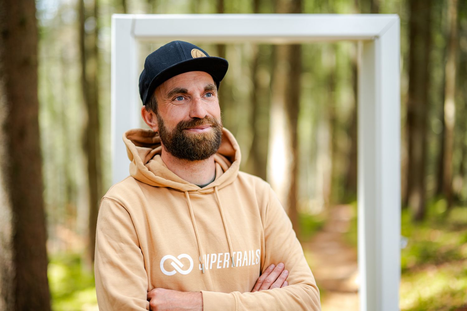 A person wearing a peach-colored hoodie and baseball cap stands in a sunlit forest setting.