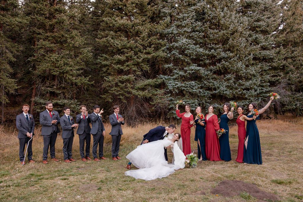 Wedding party in navy suits and burgundy bridesmaid dresses pose together in front of evergreen trees outdoors.