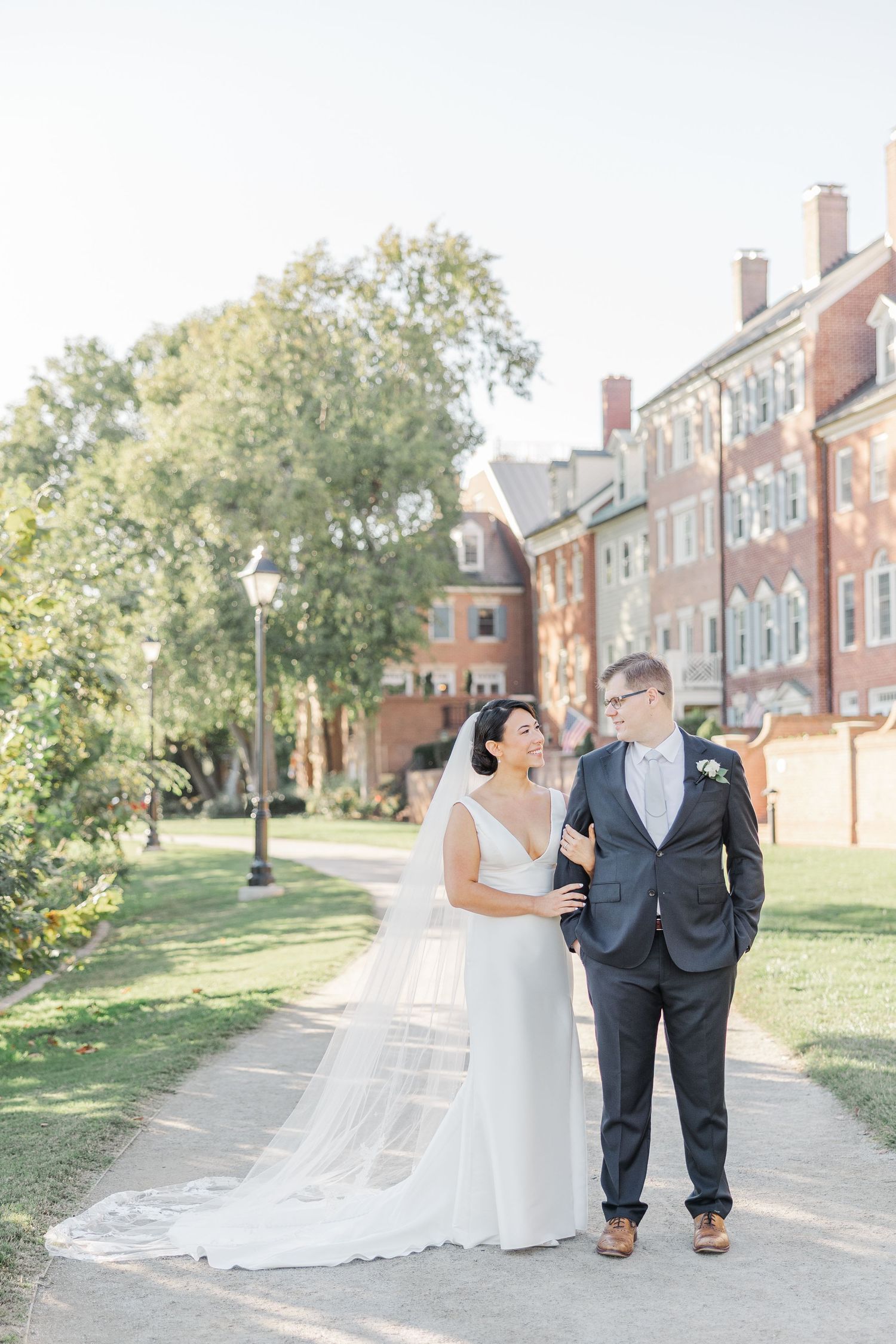 A couple in wedding attire walks together on a brick pathway surrounded by green lawns and historic brick buildings.