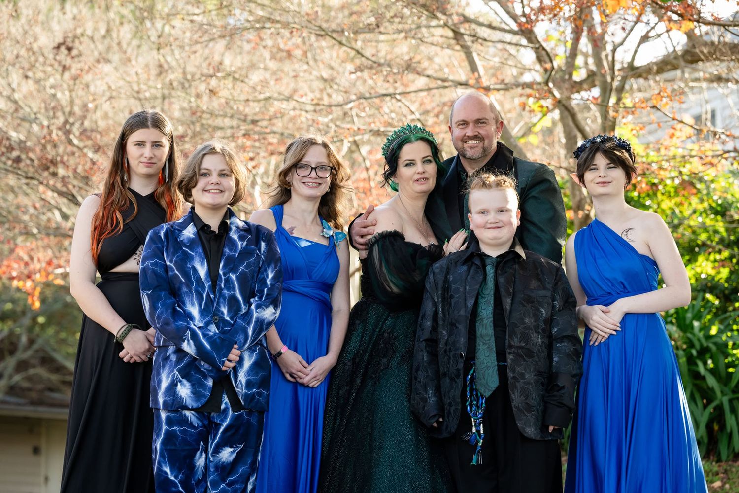 Family group portrait in formal attire, blue gowns and elegant outfits, posing outdoors with autumn foliage background.