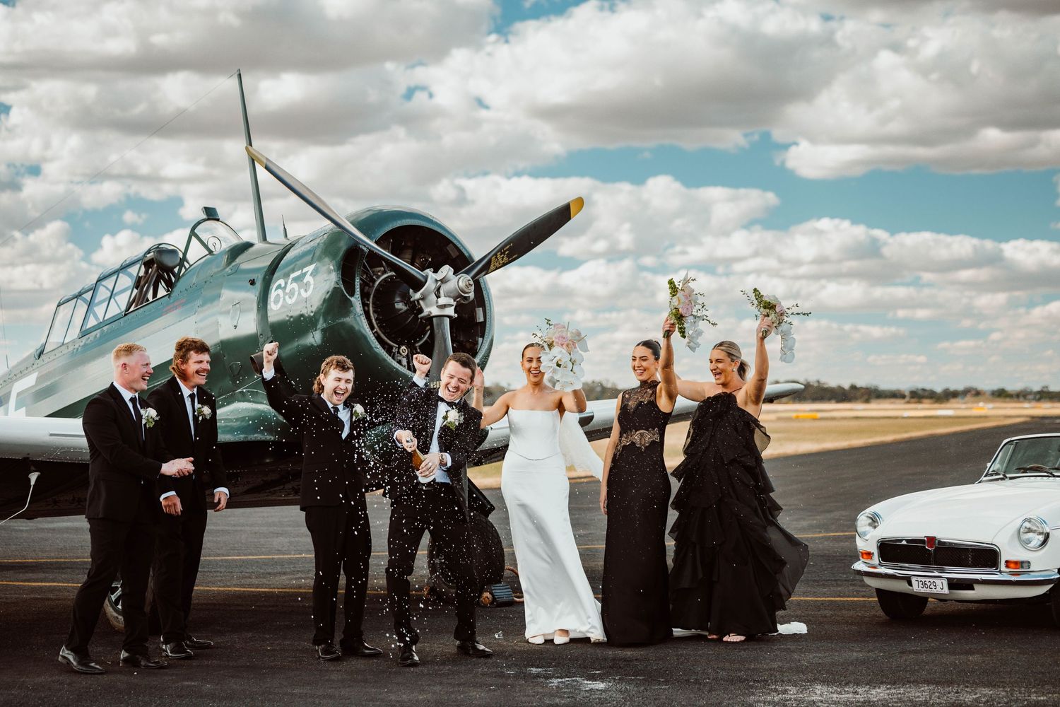 Wedding party poses joyfully by vintage aircraft on an airfield.