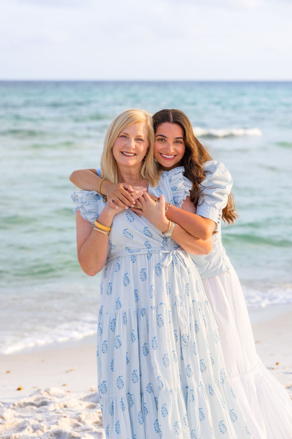 Two women in light blue and white dresses embrace while smiling on a sunny beach with turquoise ocean waves.