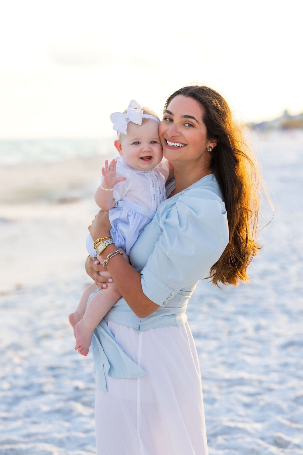 Woman in light blue top holds baby wearing white outfit and bow on sandy beach at sunset.