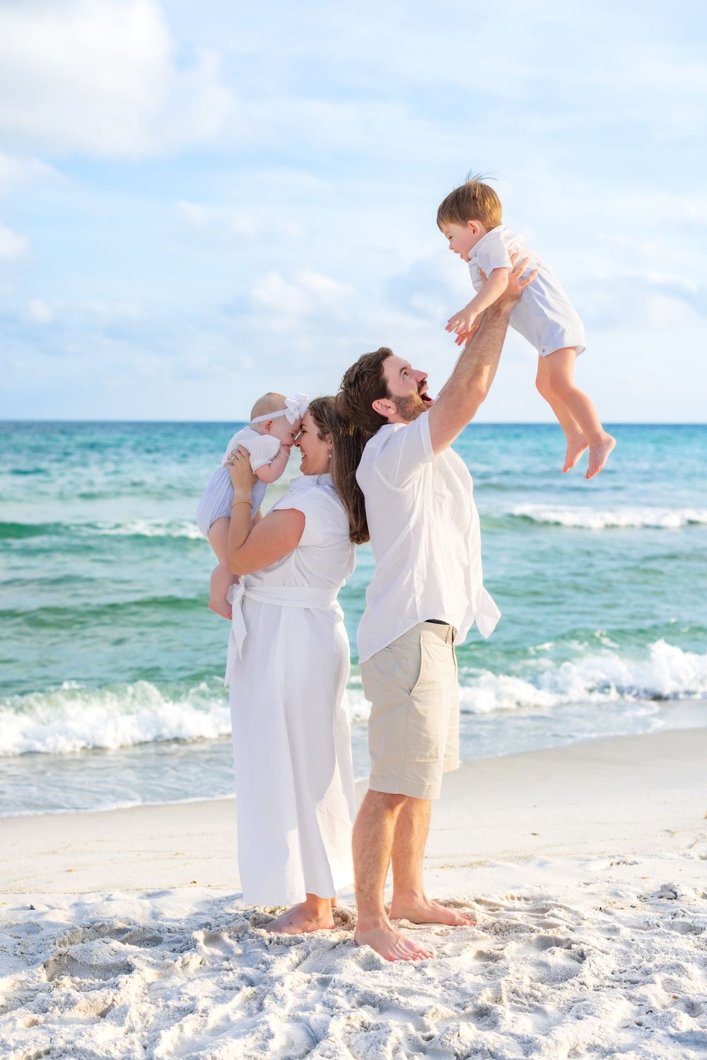 Family in white clothing playing together on a sunny beach with turquoise ocean waves in the background.