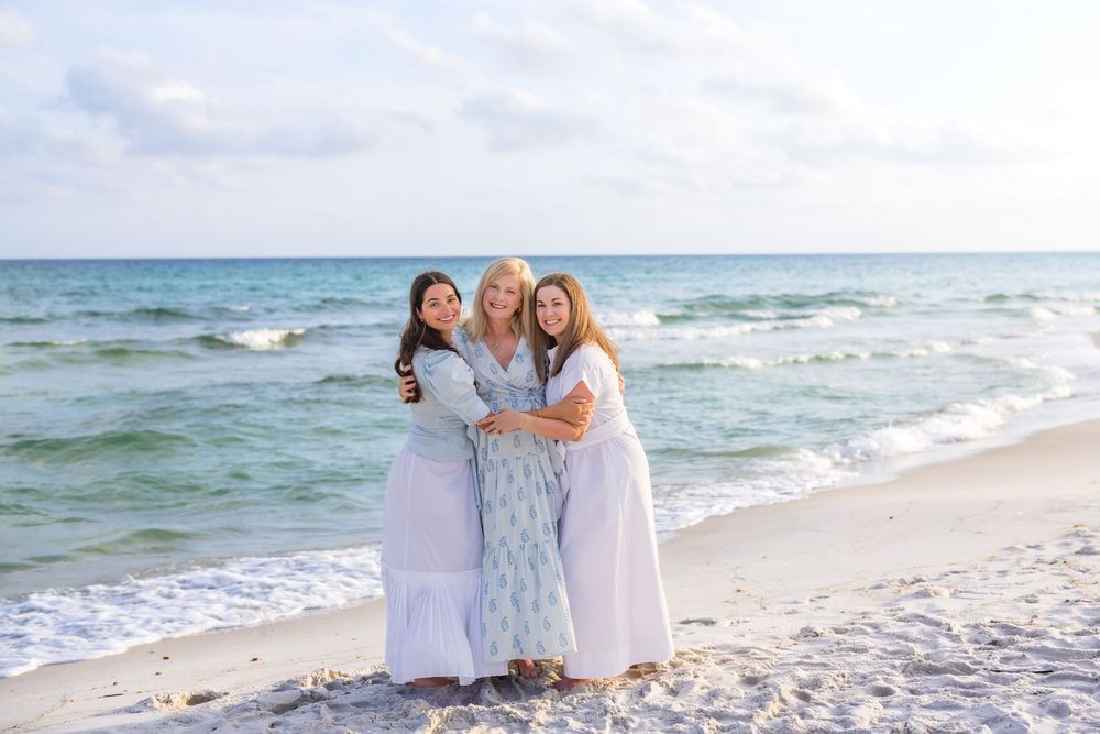 People in white outfits walk along a sandy beach with ocean waves in the background.