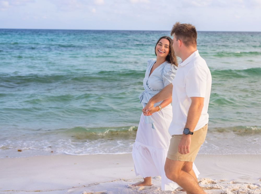 A couple in white clothing walks along a tropical beach with turquoise ocean waves in the background.