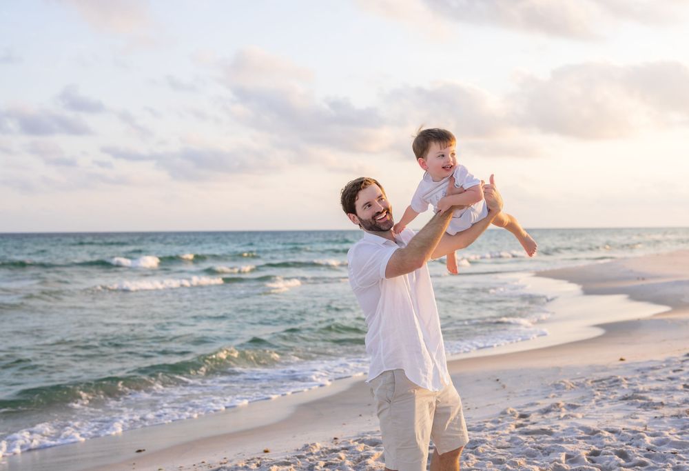 Parent lifts smiling baby in white clothing against sunset beach backdrop with ocean waves.
