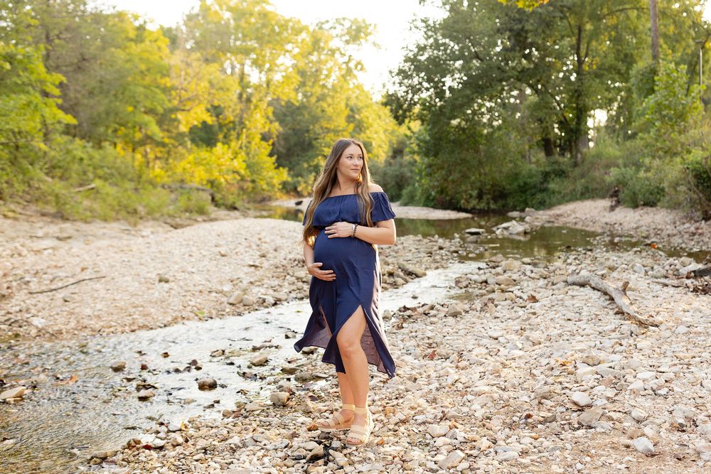 Expectant mother in navy blue dress poses on rocky riverbank during golden sunset photoshoot.