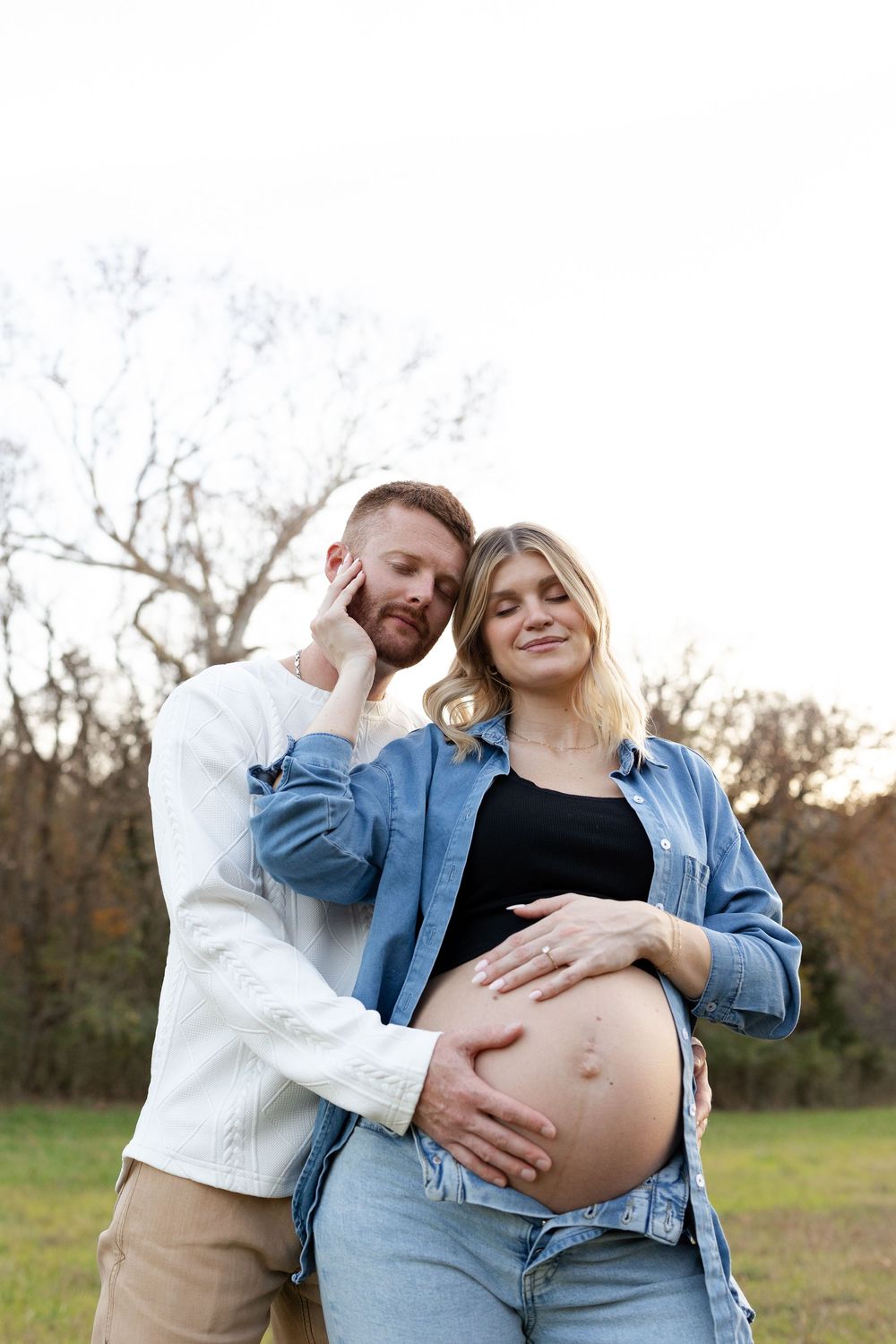 Expectant couple embraces outdoors in autumn as they touch her pregnant belly in casual attire.