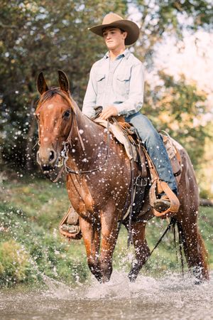 A young cowboy in a straw hat rides a sorrel horse through splashing water, with sunlit green trees behind.