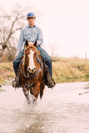 A young rider in a blue cap and denim shirt rides a sorrel horse with a white blaze, splashing through a shallow stream.